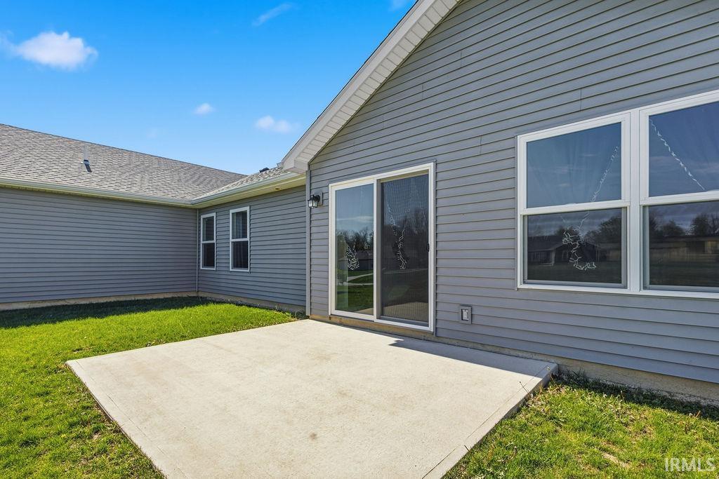 Concrete patio with a sliding glass door and multiple windows, featuring gray horizontal siding and a shingled roof