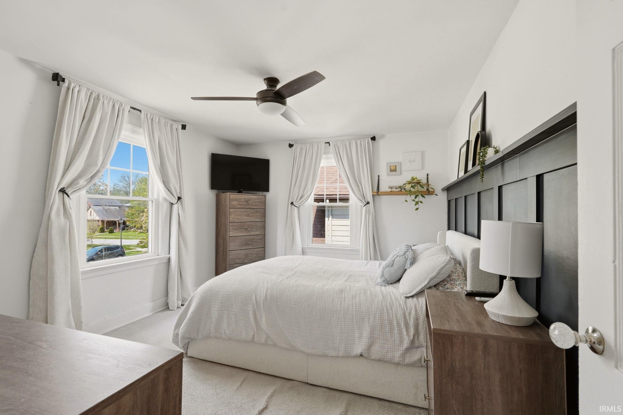 Spacious bedroom featuring multiple windows with natural light, a ceiling fan with integrated lighting, recessed wall paneling, and wood-finish flooring