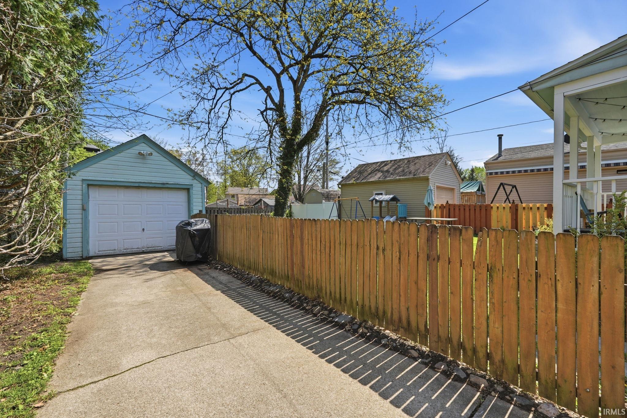 Detached garage with a white overhead door, concrete driveway, wood privacy fence, and mature landscaping