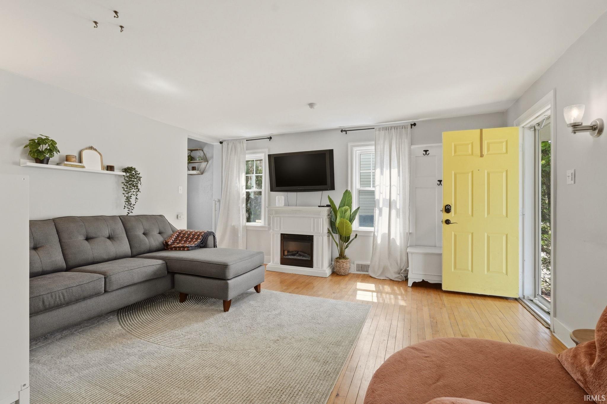 Living area featuring wood-finish flooring, a white mantel with an electric fireplace, natural light from multiple windows, and a bright paneled entry door