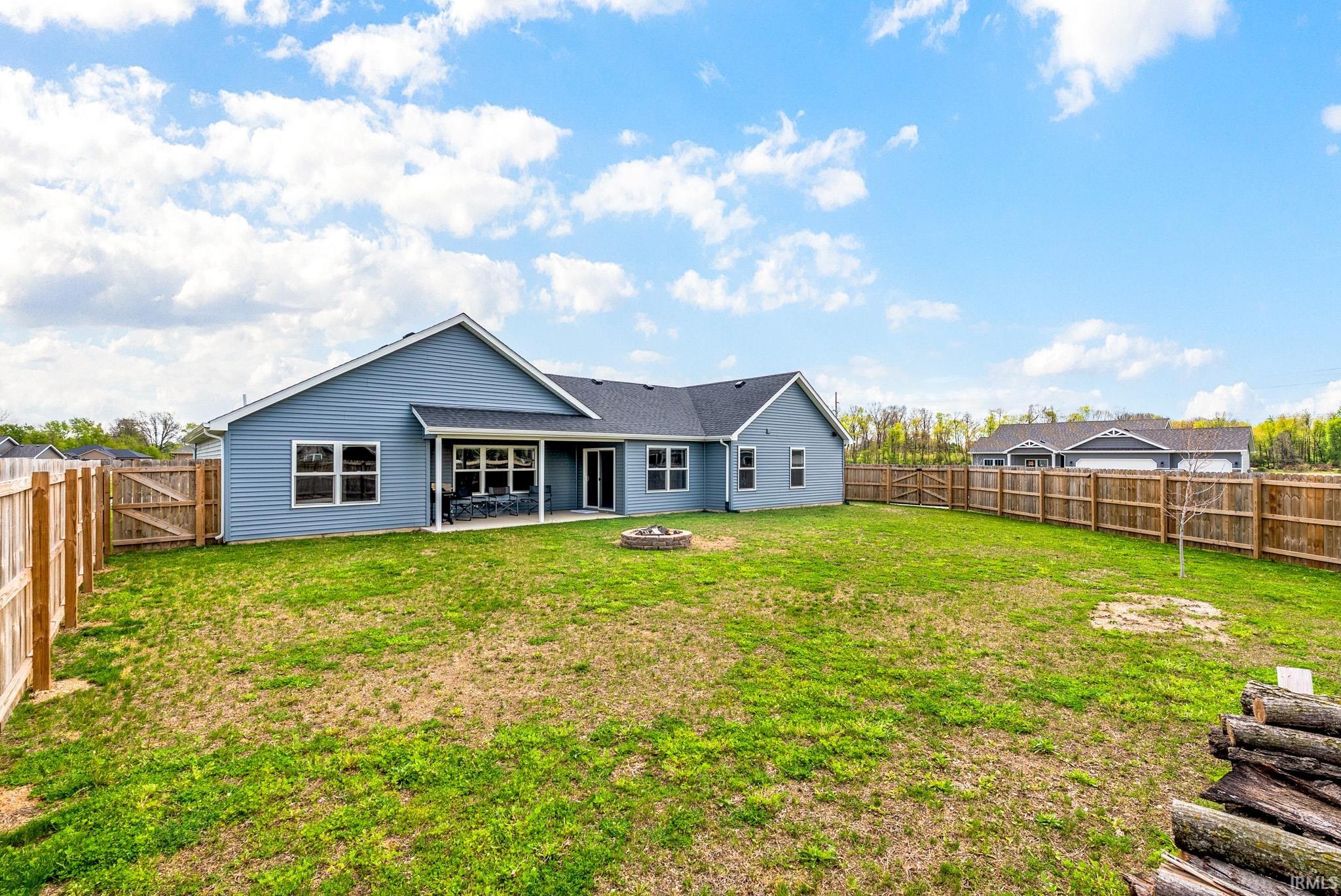 Spacious backyard featuring a light blue siding exterior, complete privacy fencing, a covered patio, and a central fire pit
