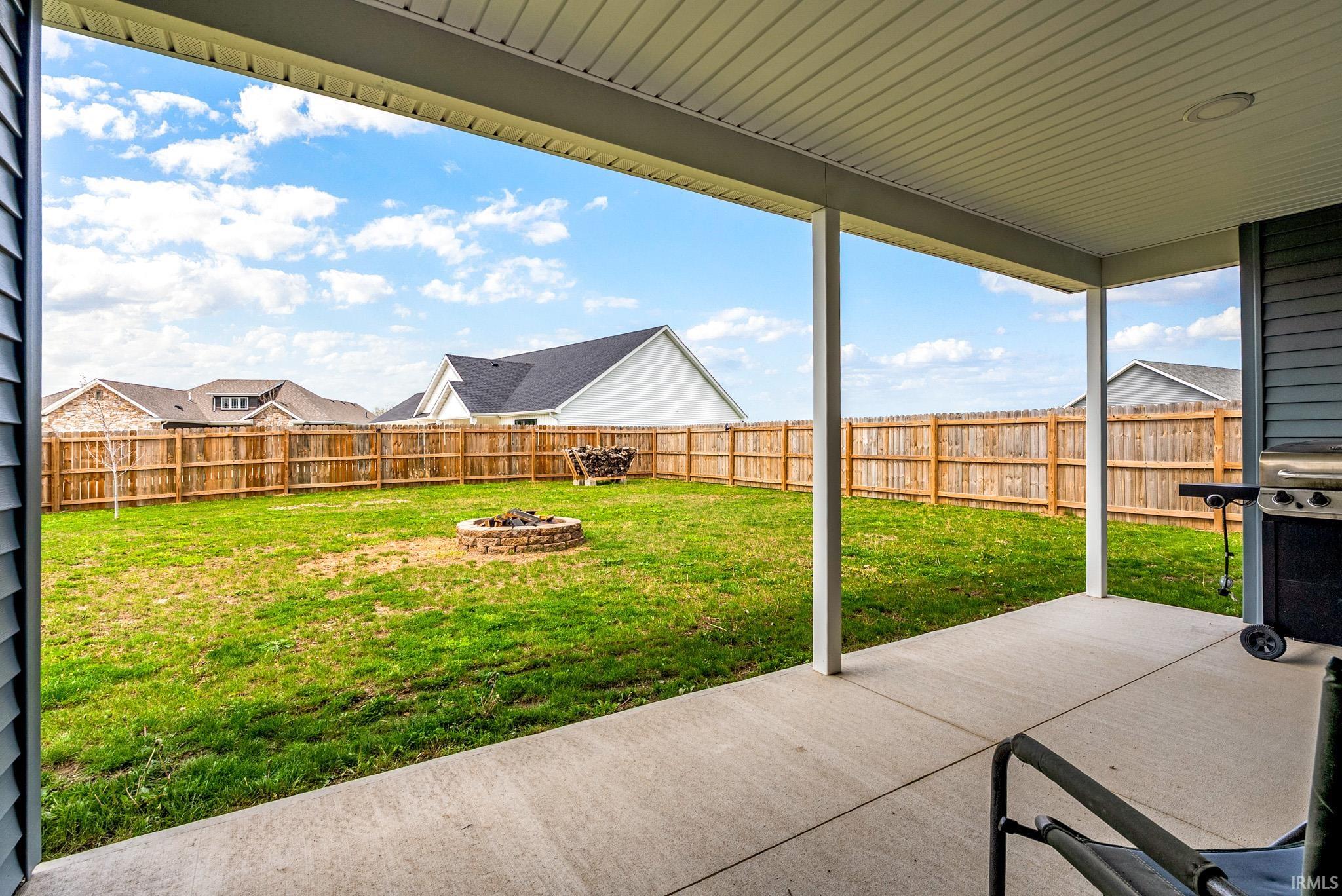 Covered patio with concrete flooring, overlooking a fenced backyard with a stone fire pit