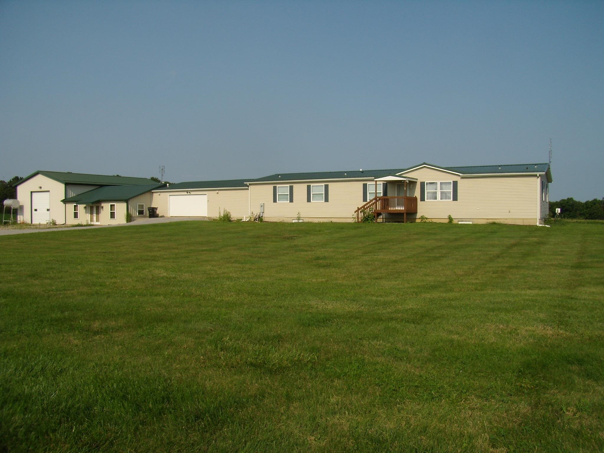 Expansive green lawn with a single-story residence featuring light-colored siding, a dark green roof, and a covered porch with wooden railing