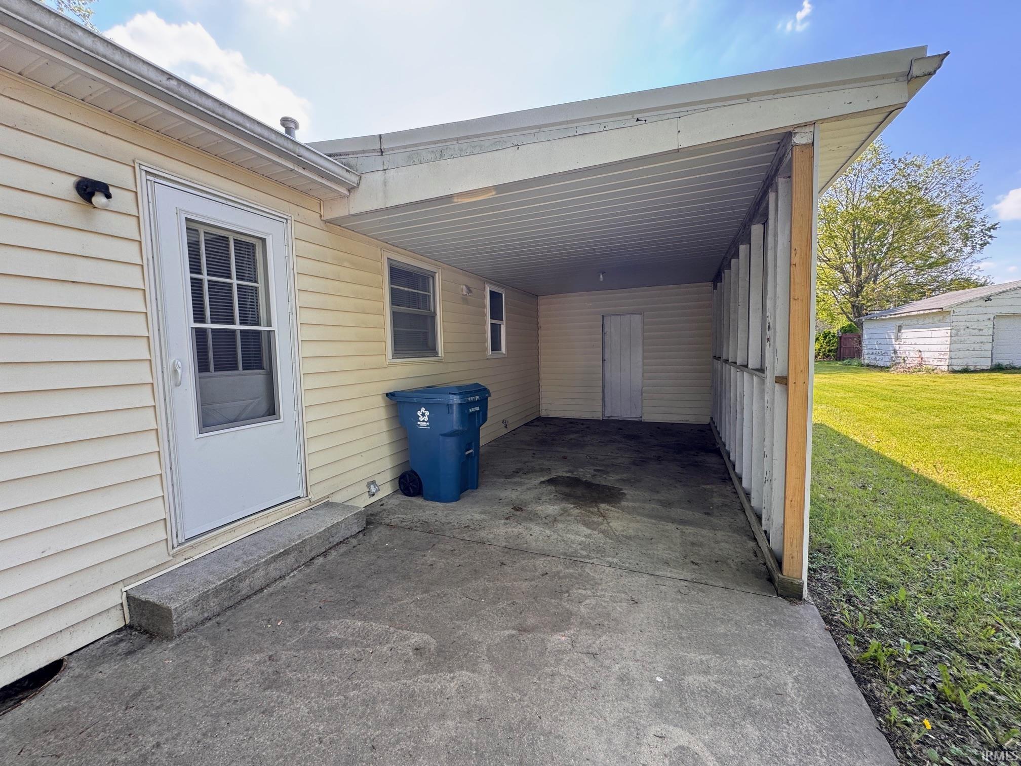 Covered carport area featuring a concrete slab, exterior access door, and storage room with a solid white door