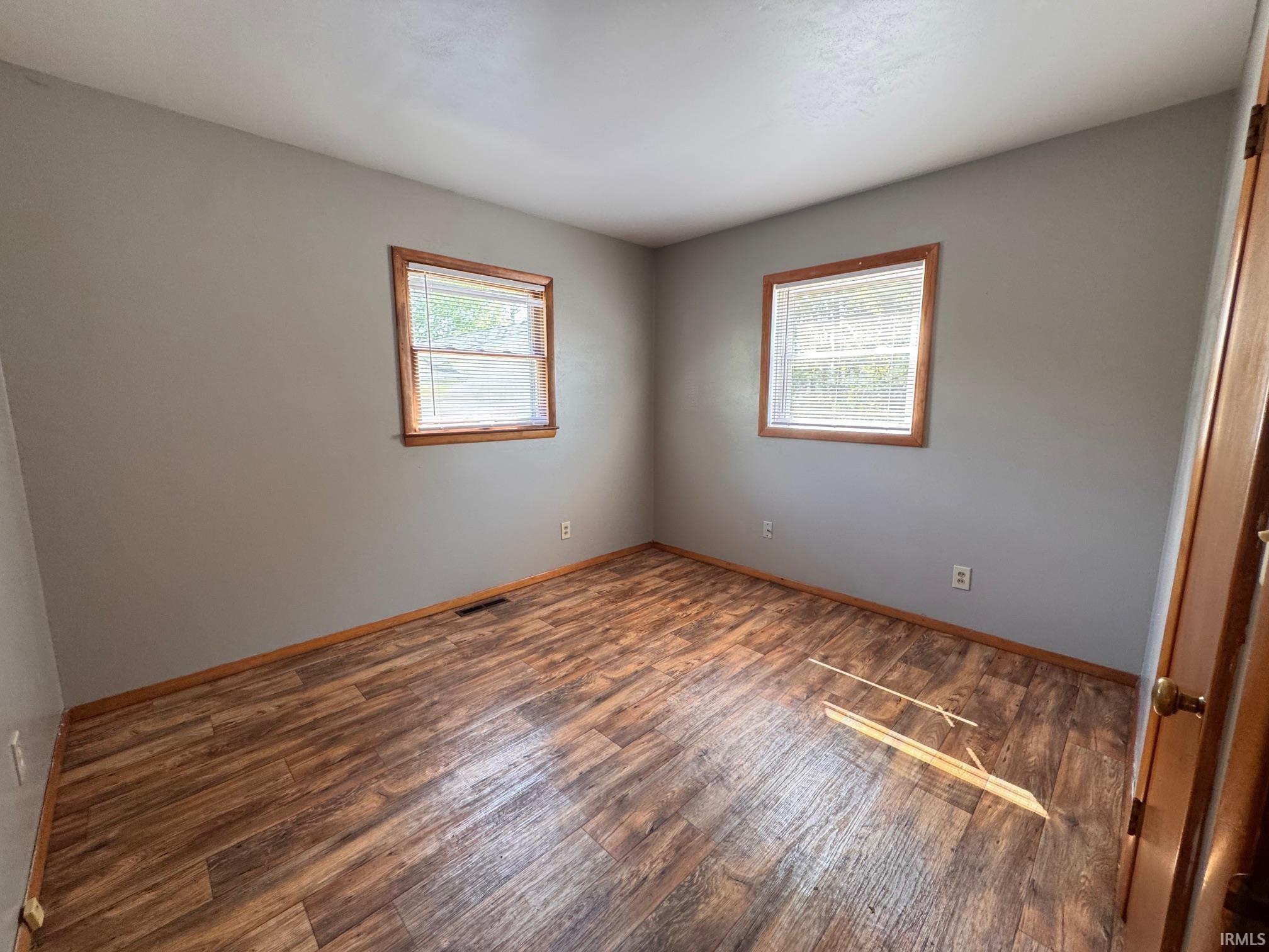 Room featuring wood-finish flooring, neutral gray walls, and two windows with wood trim