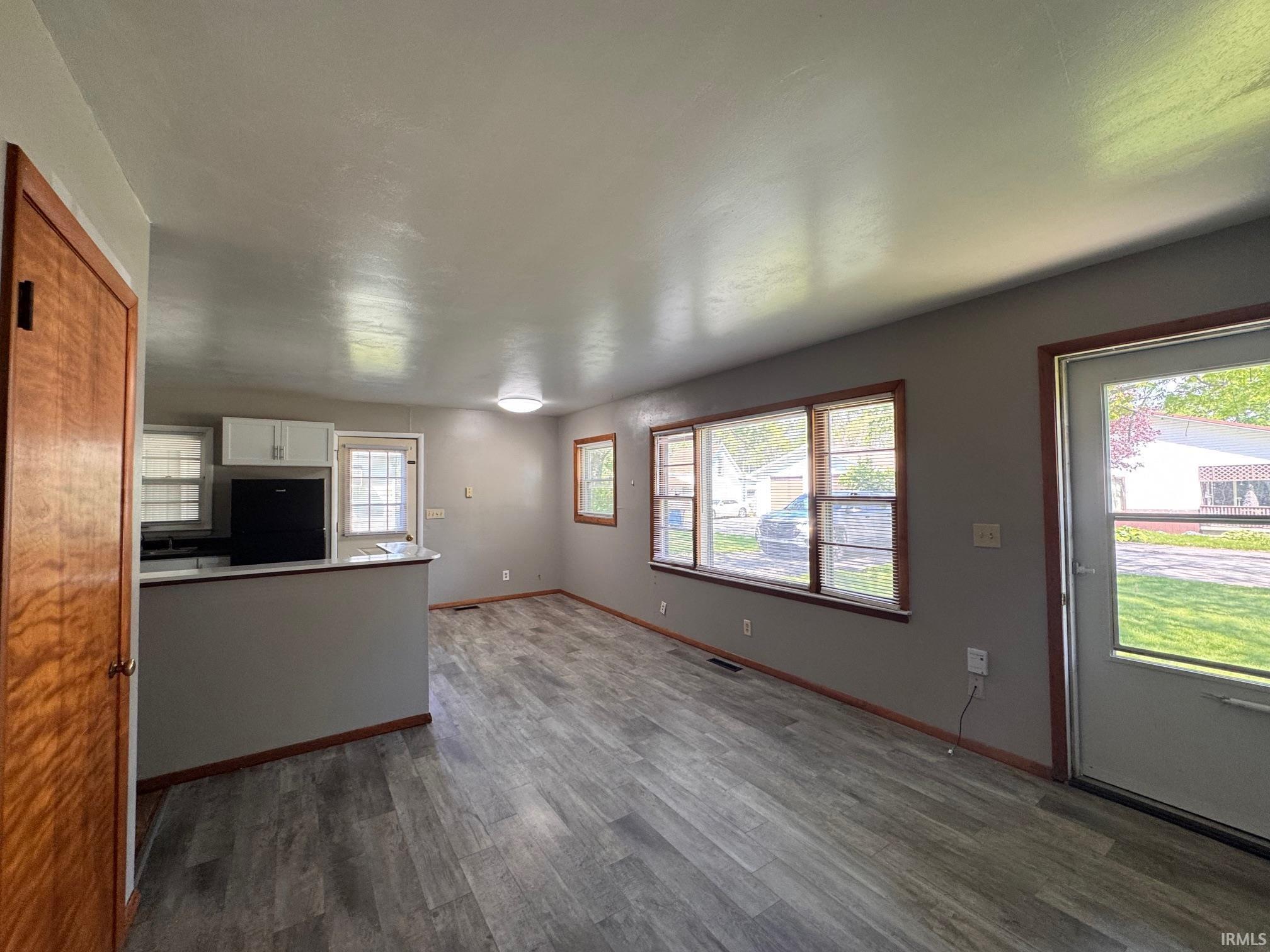 Open-concept living space featuring wood-finish flooring, multiple windows with wood trim, a kitchen pass-through, white cabinetry, and a built-in black refrigerator