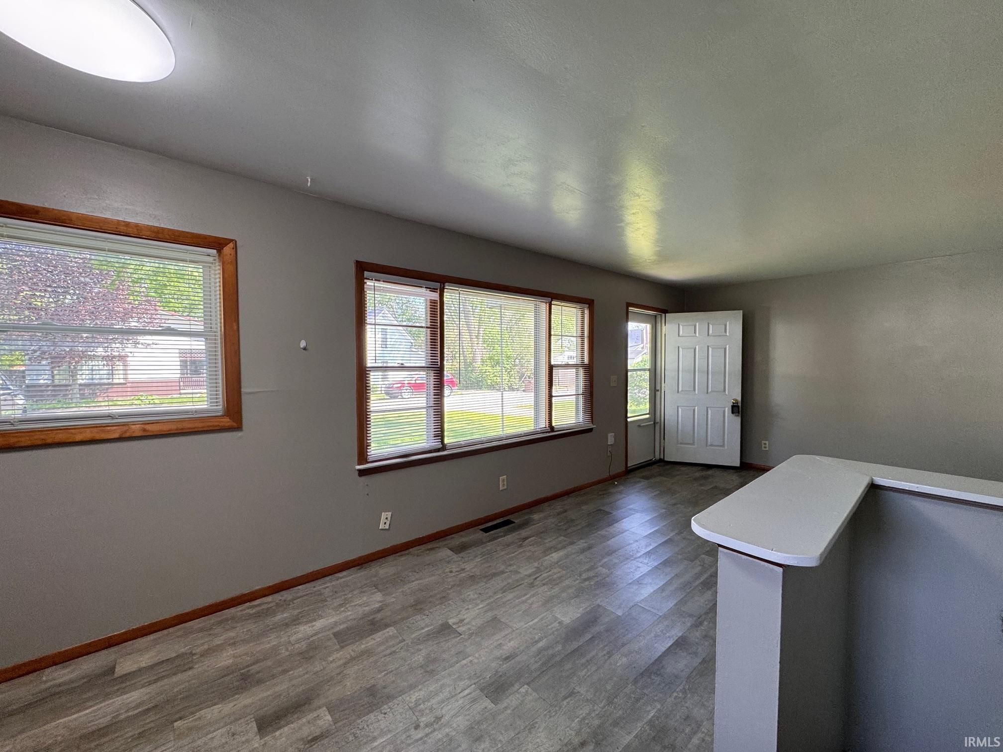 Spacious room featuring wood-finish flooring, multiple windows with wood trim, a white entry door, and a built-in half-wall with a white countertop