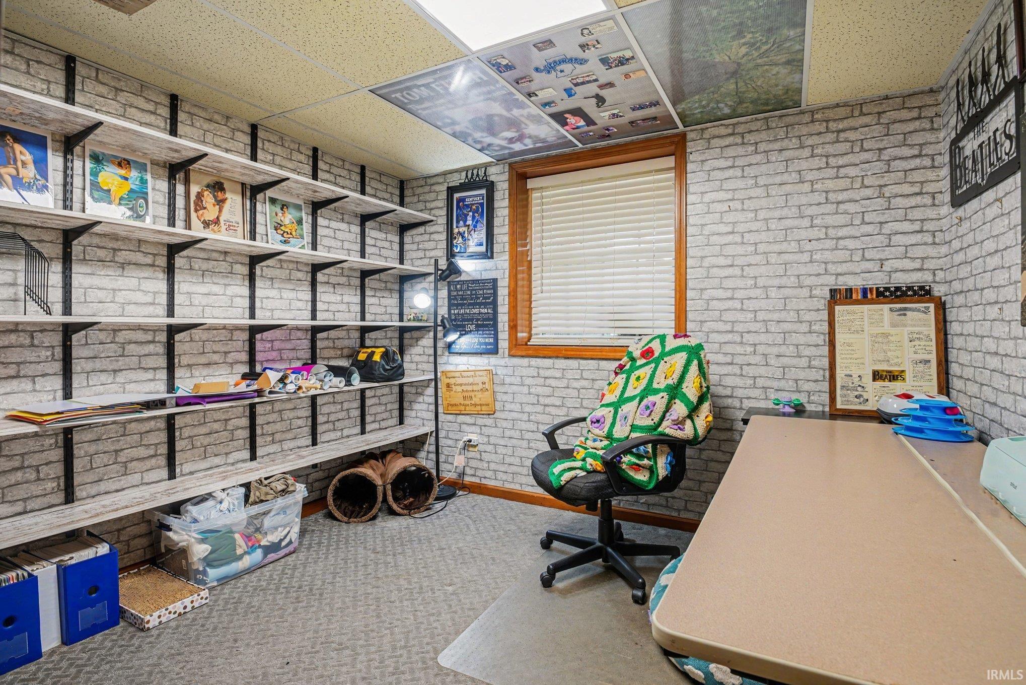 Versatile room featuring extensive wall-mounted shelving, textured brick-patterned walls, a single window with wood trim, and a drop ceiling
