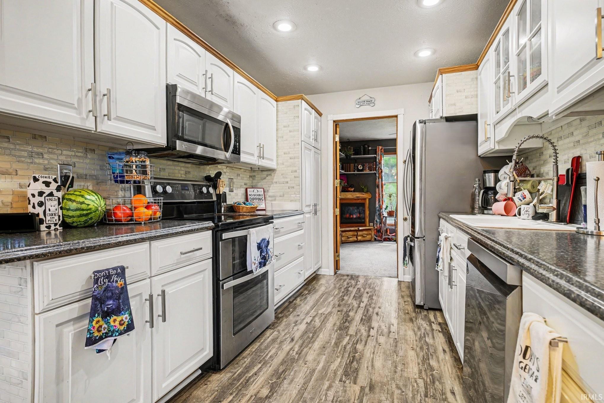 Kitchen featuring white cabinetry, dark countertops, a mosaic tile backsplash, stainless steel appliances, and wood-finish flooring