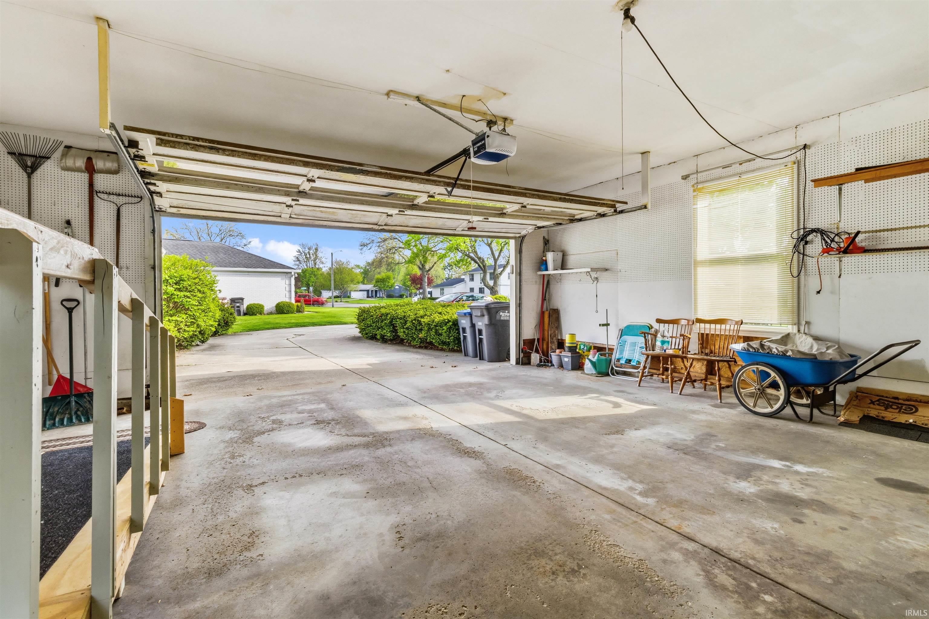 Attached garage with an open overhead door, concrete flooring, window with blinds, and built-in pegboard storage