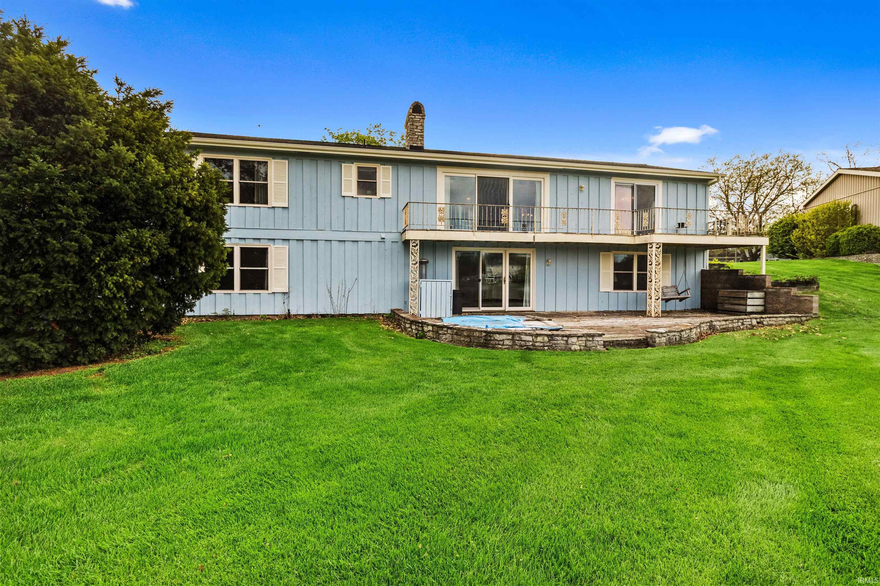 Two-story property featuring a light blue exterior, upper-level balcony with metal railings, and a lower-level stone patio