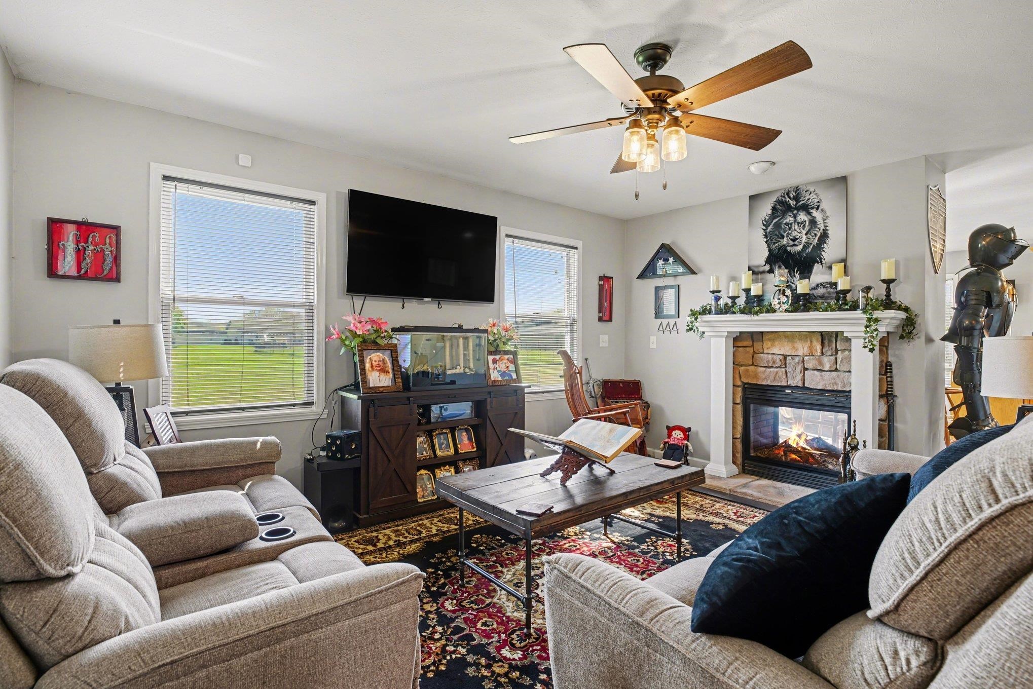 Living area featuring a stone-surround fireplace with a white mantel, ceiling fan with integrated lighting, and multiple windows