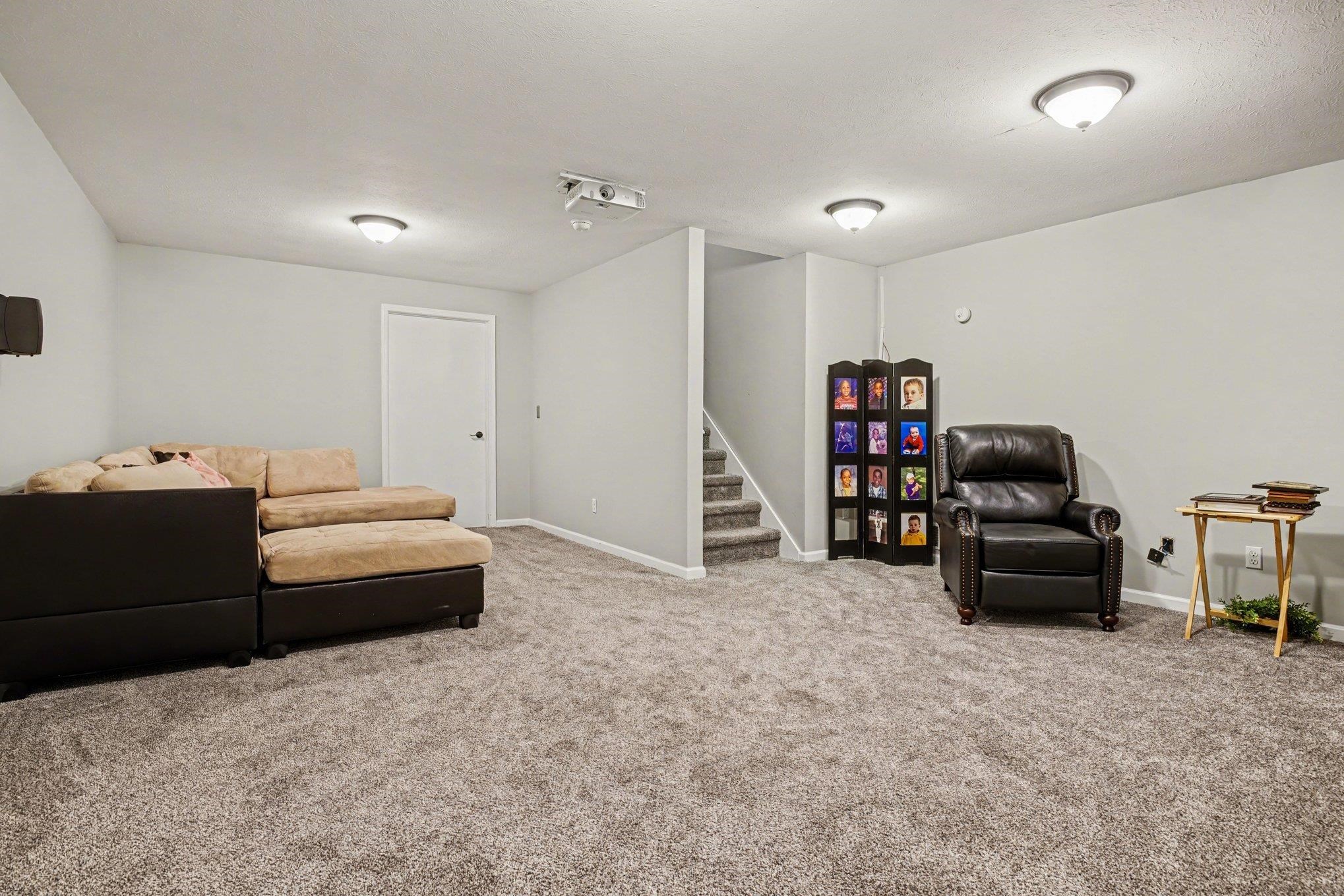 Spacious carpeted room featuring a ceiling-mounted projector, multiple ceiling light fixtures, a white interior door, and a carpeted staircase