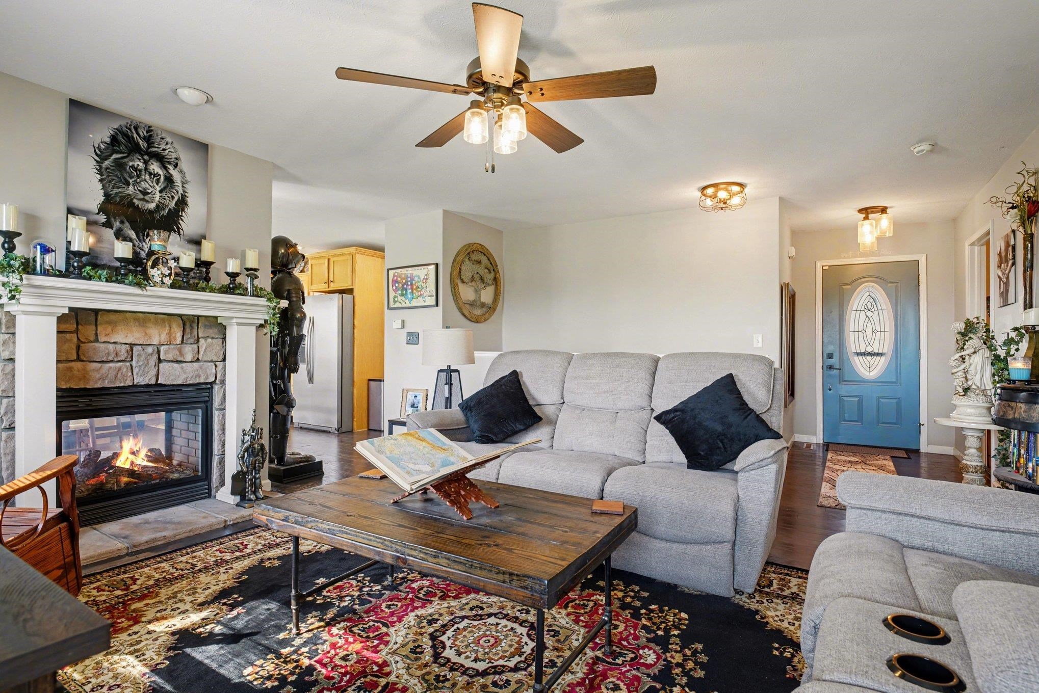 Living area featuring a stone-clad fireplace with a white mantel, wood-finish flooring, and a ceiling fan with integrated lighting
