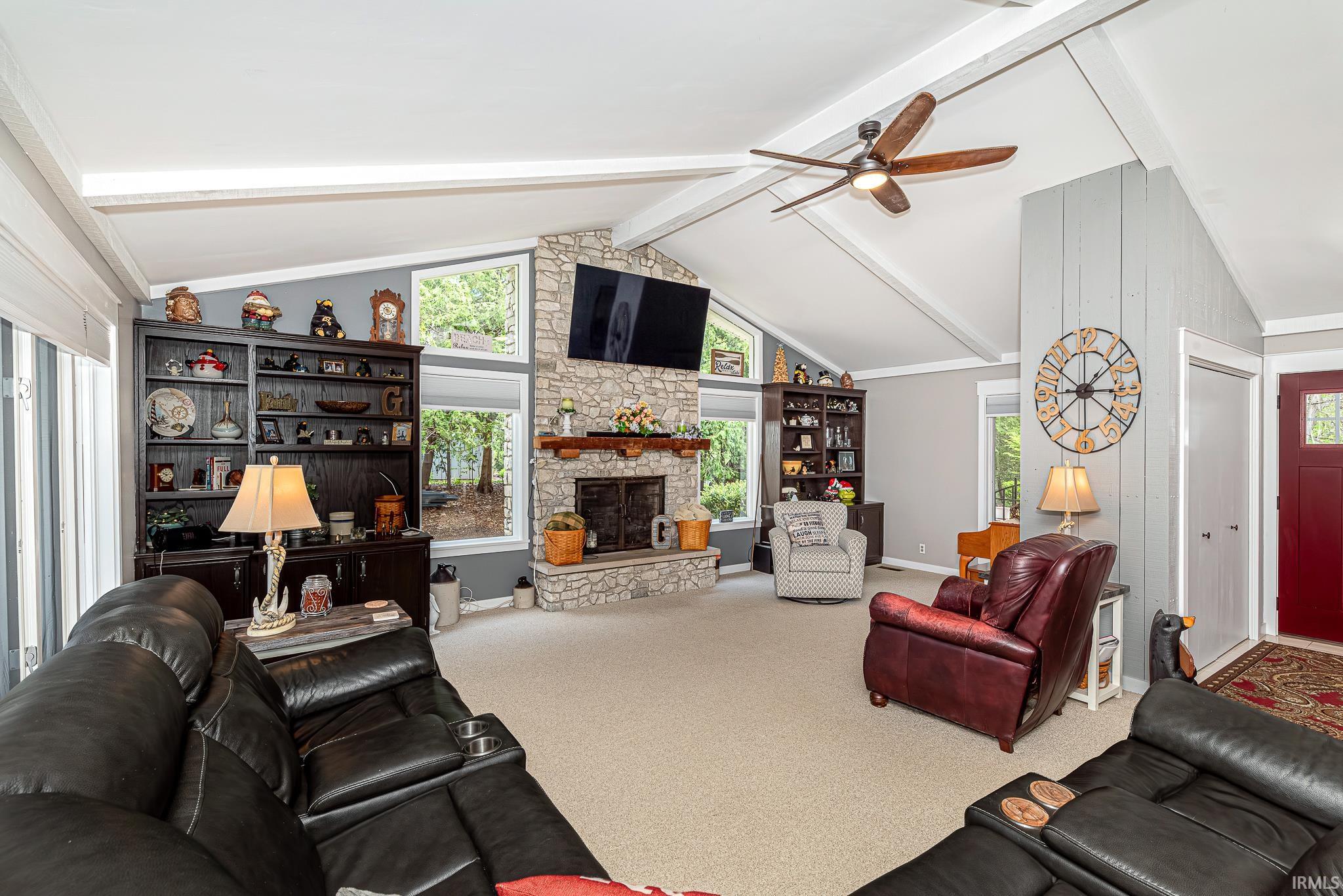 Spacious living area featuring a stone fireplace with a wood mantle, vaulted ceilings with exposed beams, and multiple large windows