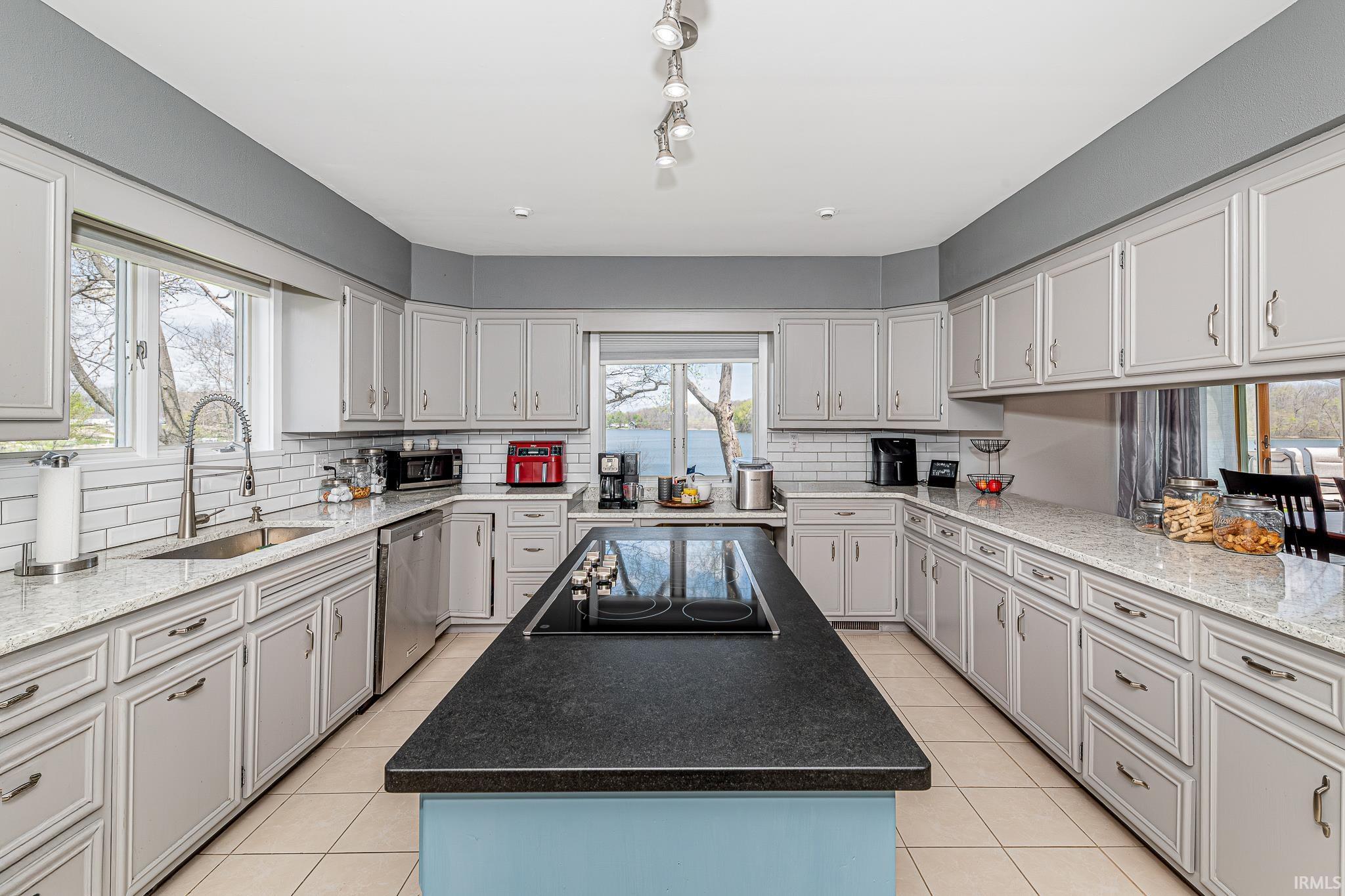 Spacious kitchen featuring a central island with a built-in cooktop, extensive cabinetry with light gray finishes, light-toned countertops, a stainless steel sink with a gooseneck faucet, and white subway tile backsplash