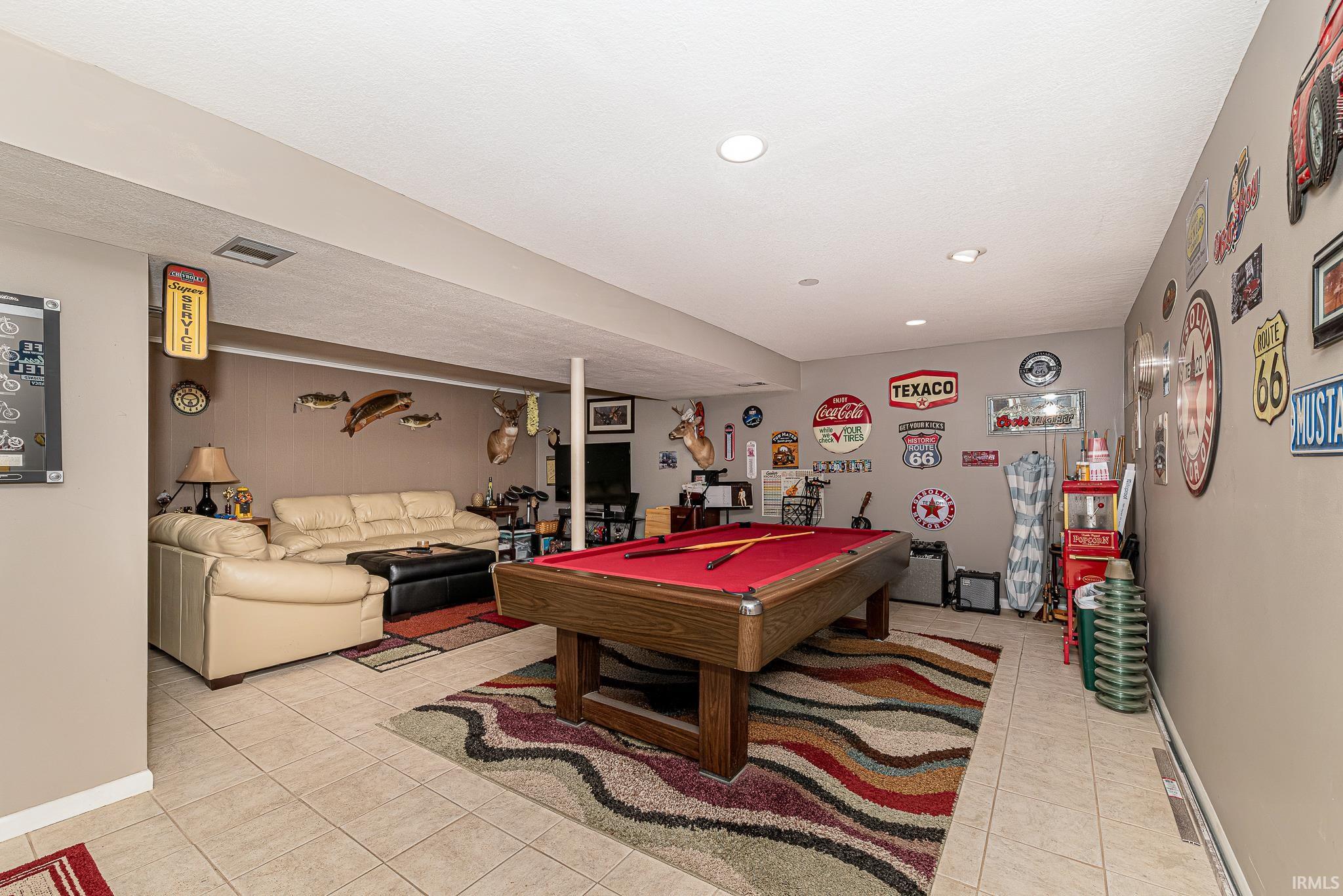 Spacious recreation room featuring a billiards table with a red felt surface, recessed ceiling lighting, light-toned tile flooring, and a dedicated lounge area