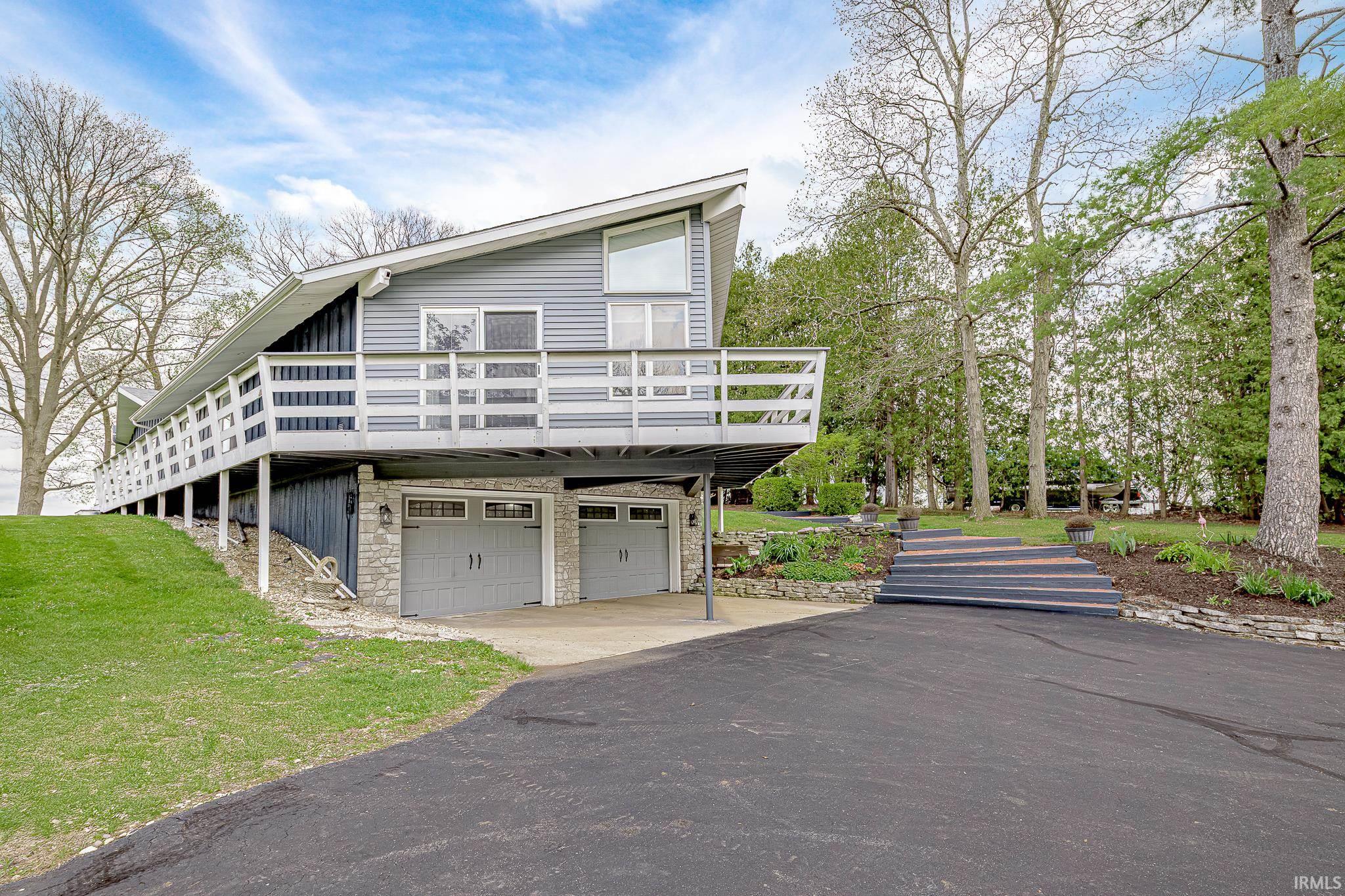 Contemporary architectural style residence featuring a two-car garage with stone accents, an extended upper-level deck, a sloped asphalt driveway, and a terraced landscaped front yard with stone retaining walls