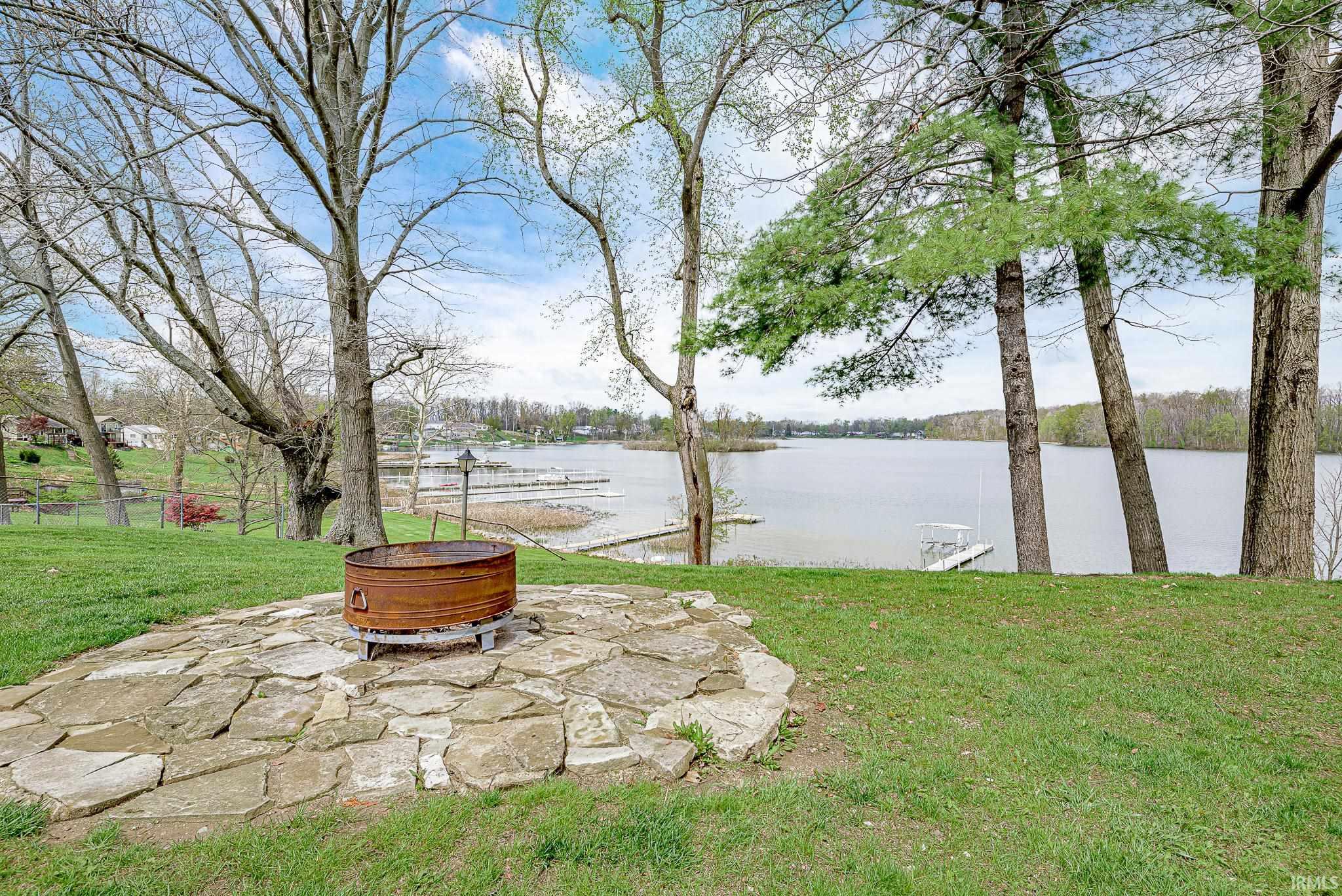 Stone paver patio featuring a central fire pit, surrounded by a grassy lawn and mature trees, with a lake and distant shoreline visible