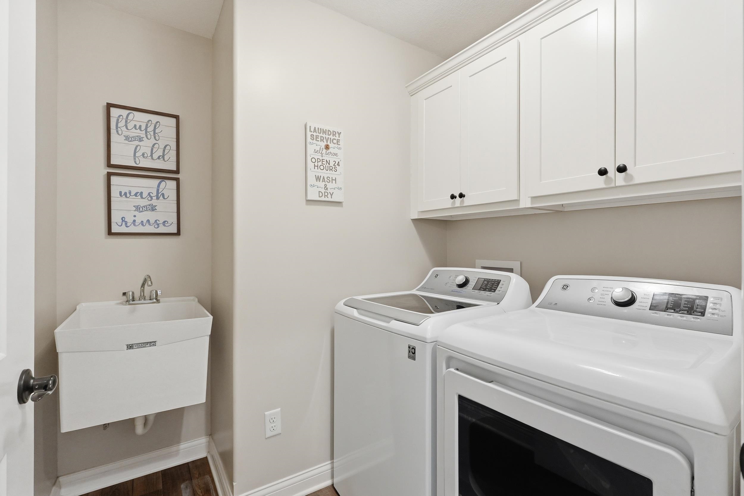 Dedicated laundry room featuring upper white shaker cabinetry with dark hardware, a utility sink with stainless steel faucet, and wood-finish flooring