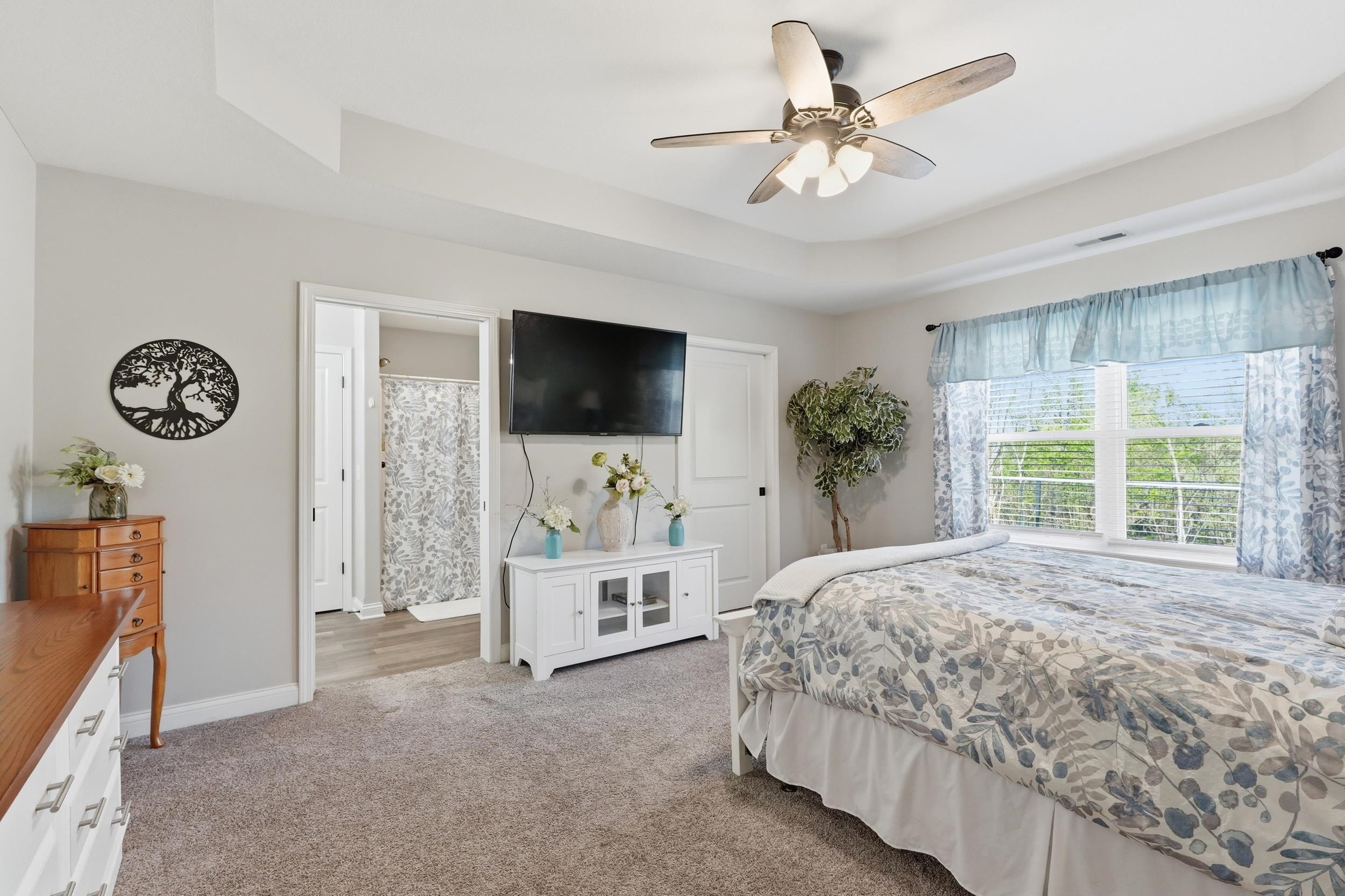 Carpeted bedroom featuring a tray ceiling with a ceiling fan, a window with blinds, and an attached bathroom with wood-finish flooring