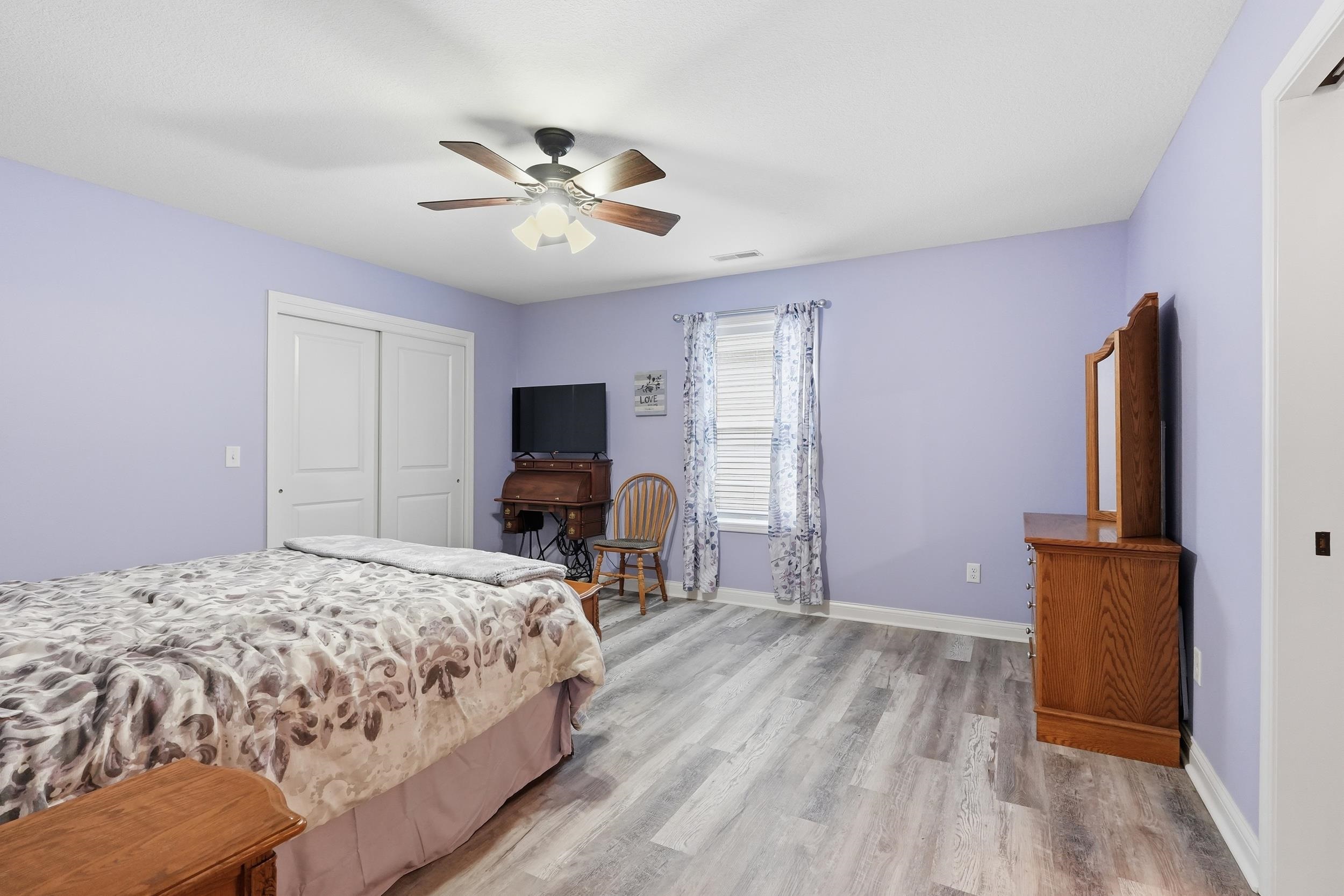 Spacious room featuring wood-finish flooring, a ceiling fan with integrated lighting, a window with blinds, and white bi-fold closet doors