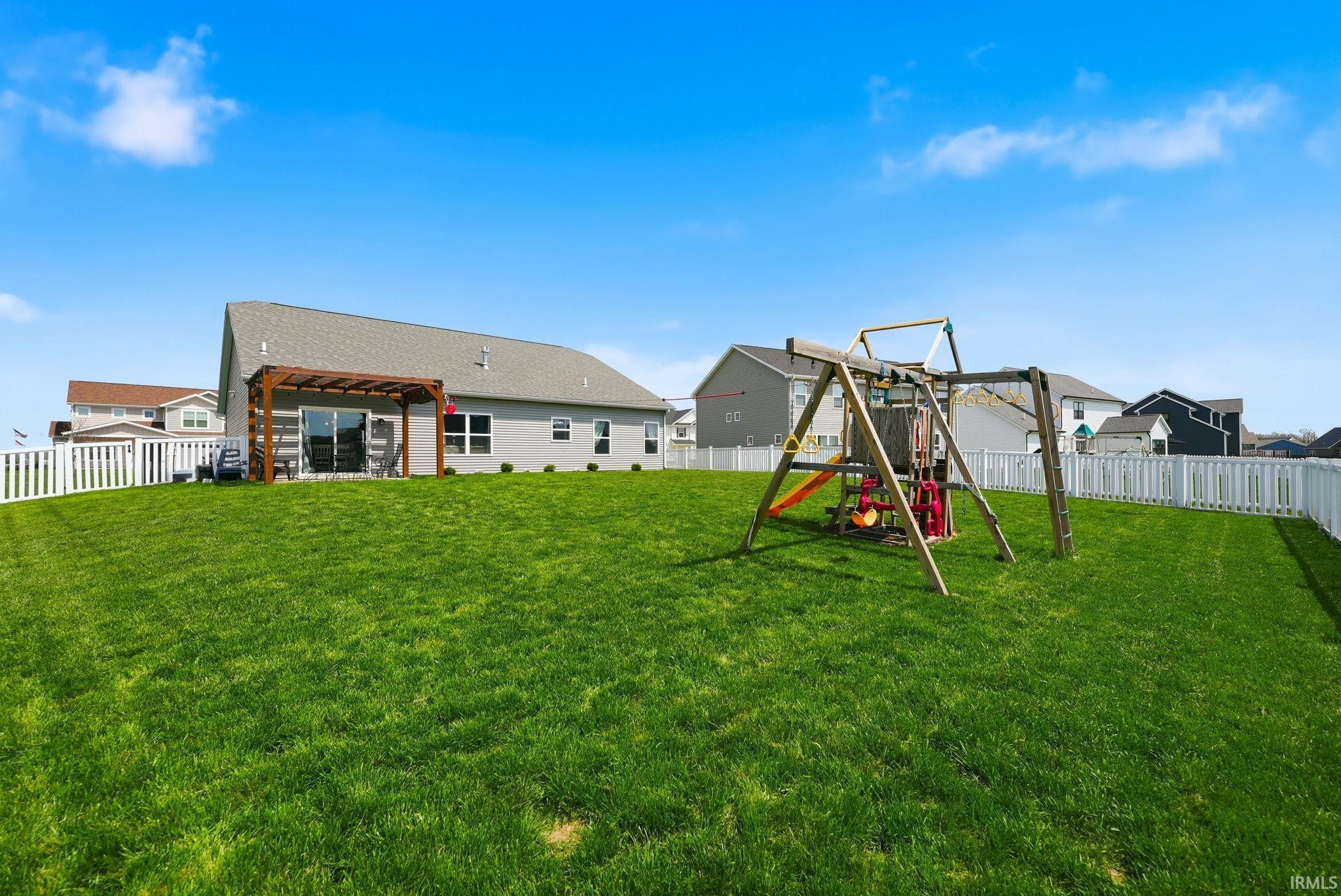 Expansive green lawn with a white picket fence, featuring a grey siding home with a wooden pergola over the patio