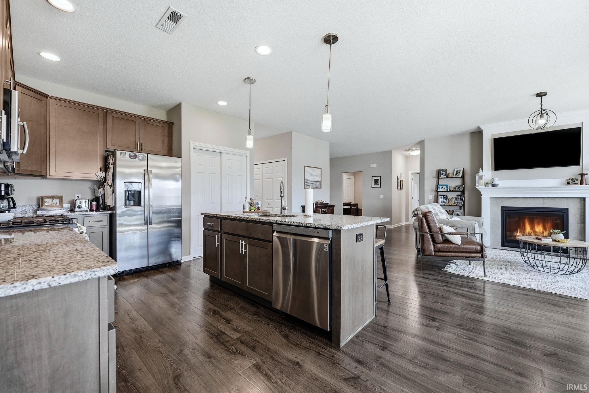 Open-concept living area featuring wood-finish flooring, a kitchen island with integrated sink, stainless steel appliances, and a fireplace with a white mantel