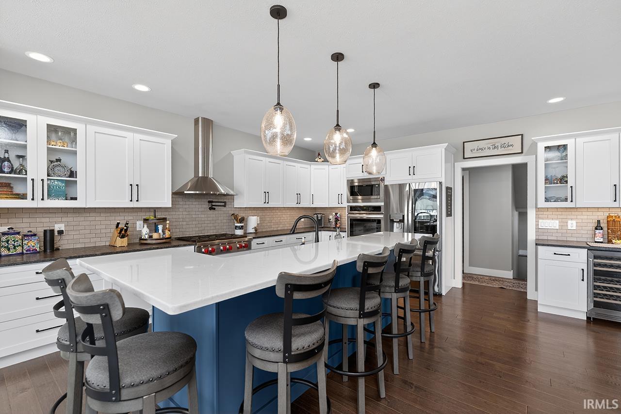 Spacious kitchen featuring an expansive center island with a white countertop and blue base, white cabinetry with dark hardware, stainless steel appliances, a tile backsplash, and wood-finish flooring