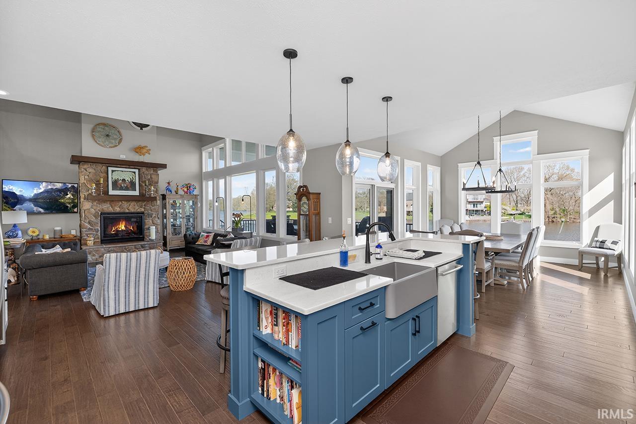 Open concept interior featuring wood-finish flooring, a kitchen island with a farmhouse sink and blue cabinetry, a stone-clad fireplace, and vaulted ceilings with large windows