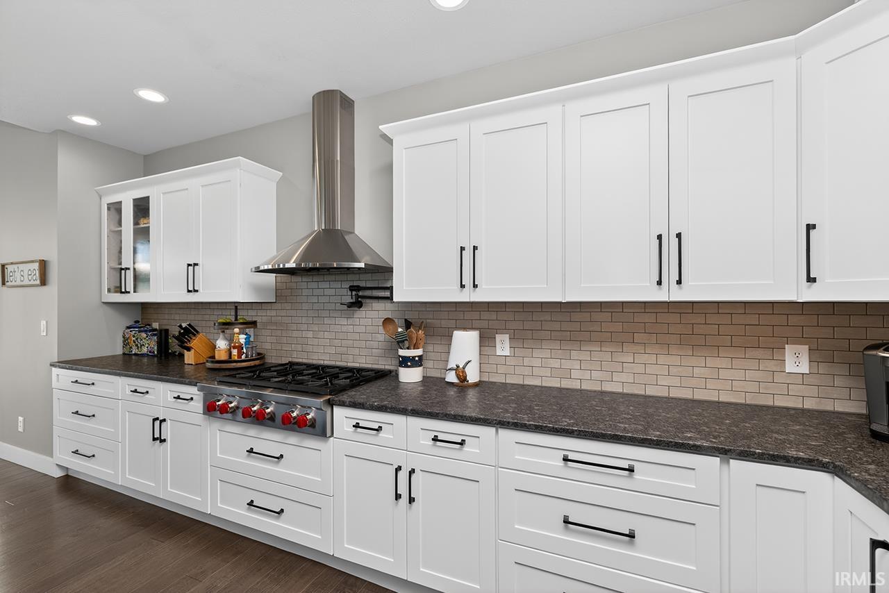 White shaker cabinetry with dark hardware, stone-finish countertops, stainless steel range with hood, glass tile backsplash, and recessed ceiling lighting
