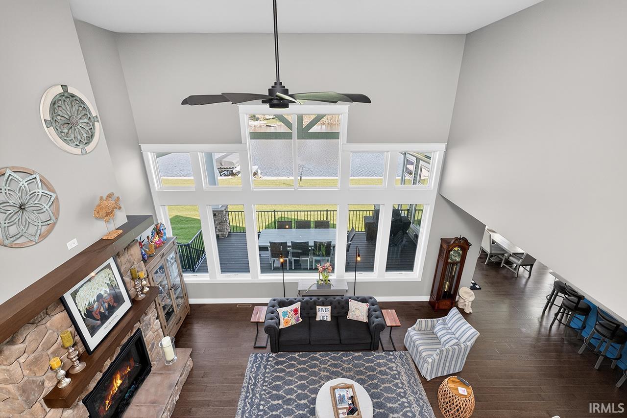 Vaulted living area featuring a stone fireplace with a wood mantle, an array of large picture windows, wood-finish flooring, and a ceiling fan