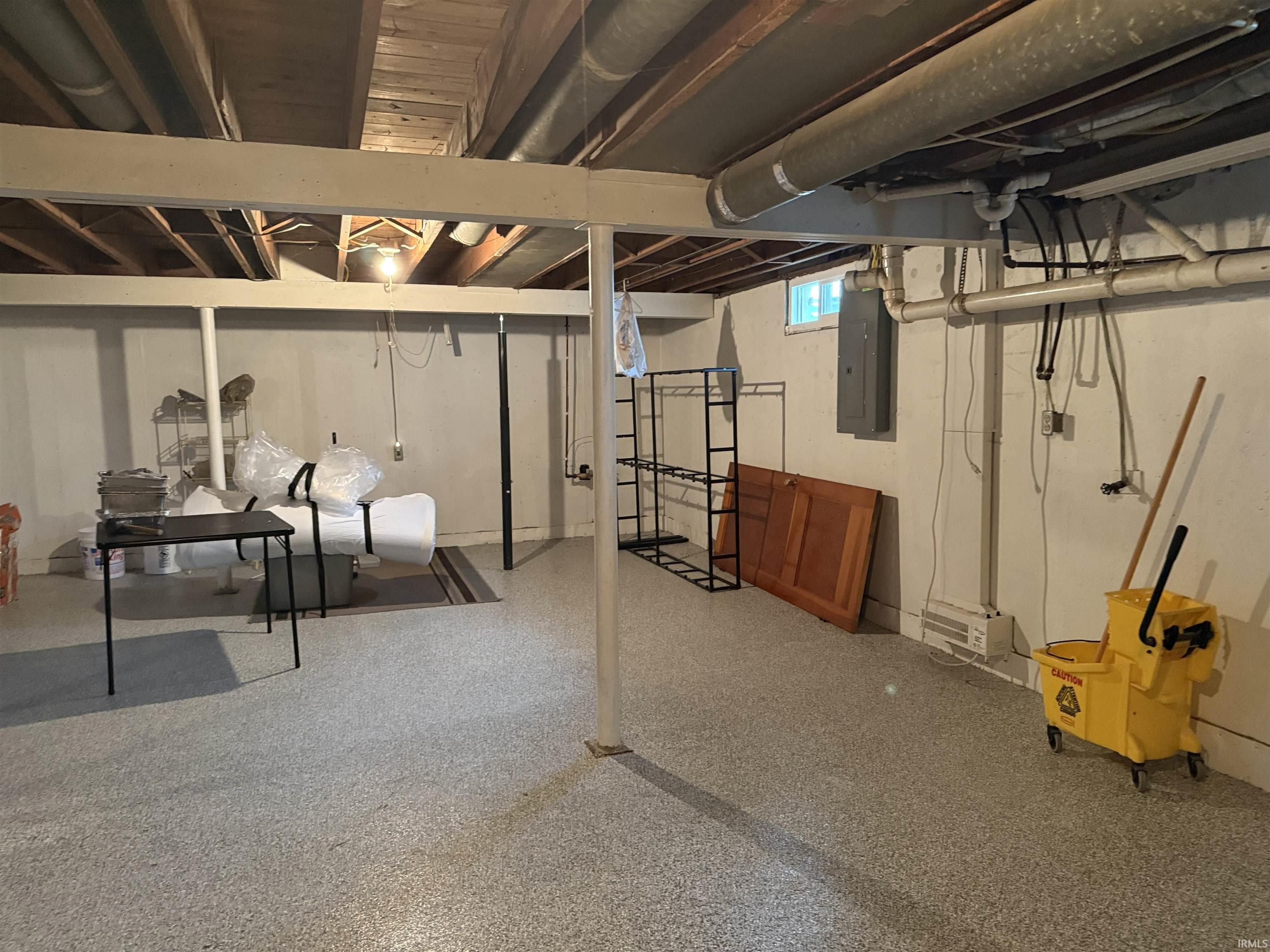 Unfinished basement with exposed ceiling joists and support beams, featuring a speckled concrete floor, painted concrete walls, and visible utility infrastructure