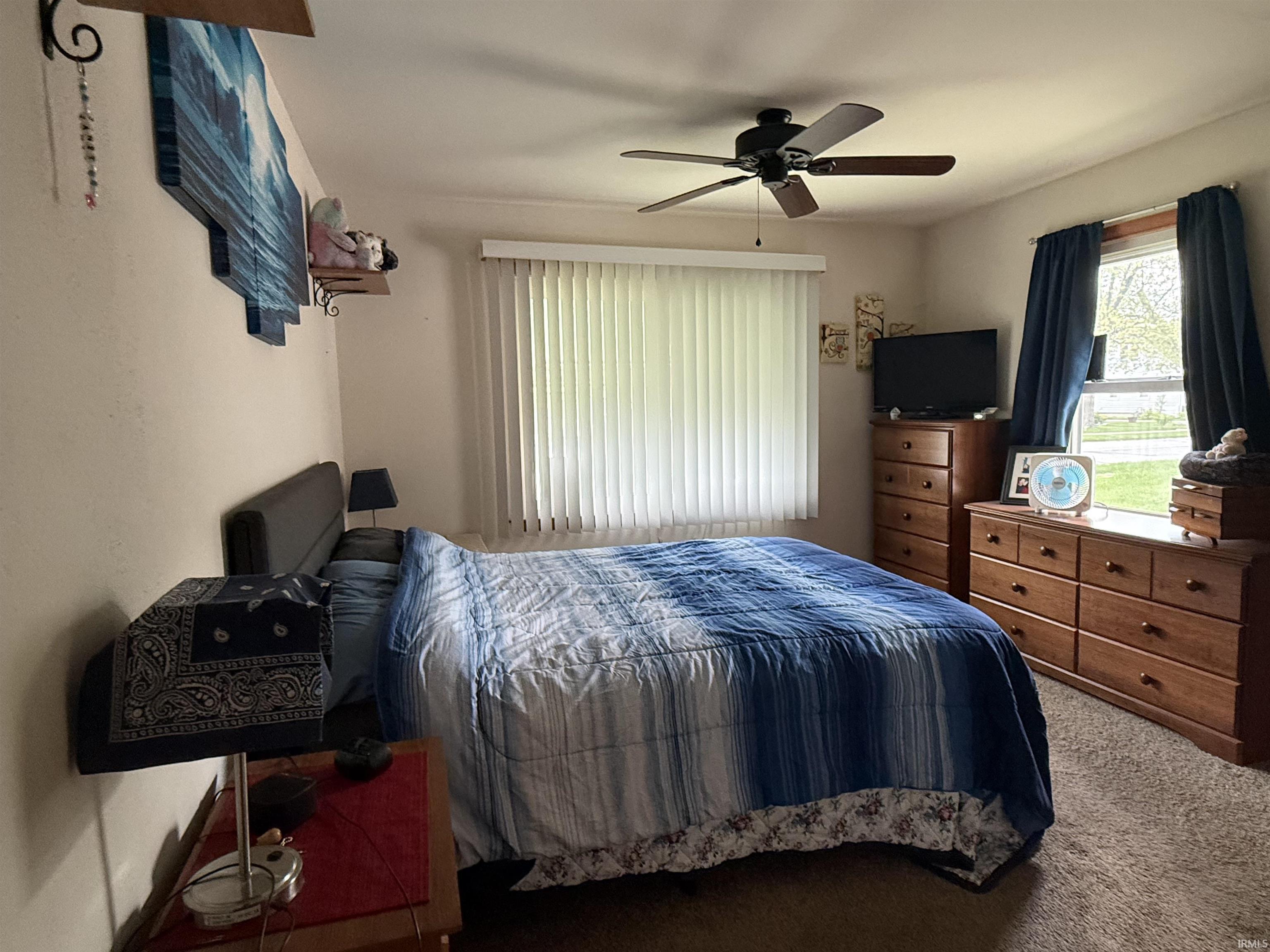 Carpeted room with a ceiling fan, window with vertical blinds, and a second window with blue curtains