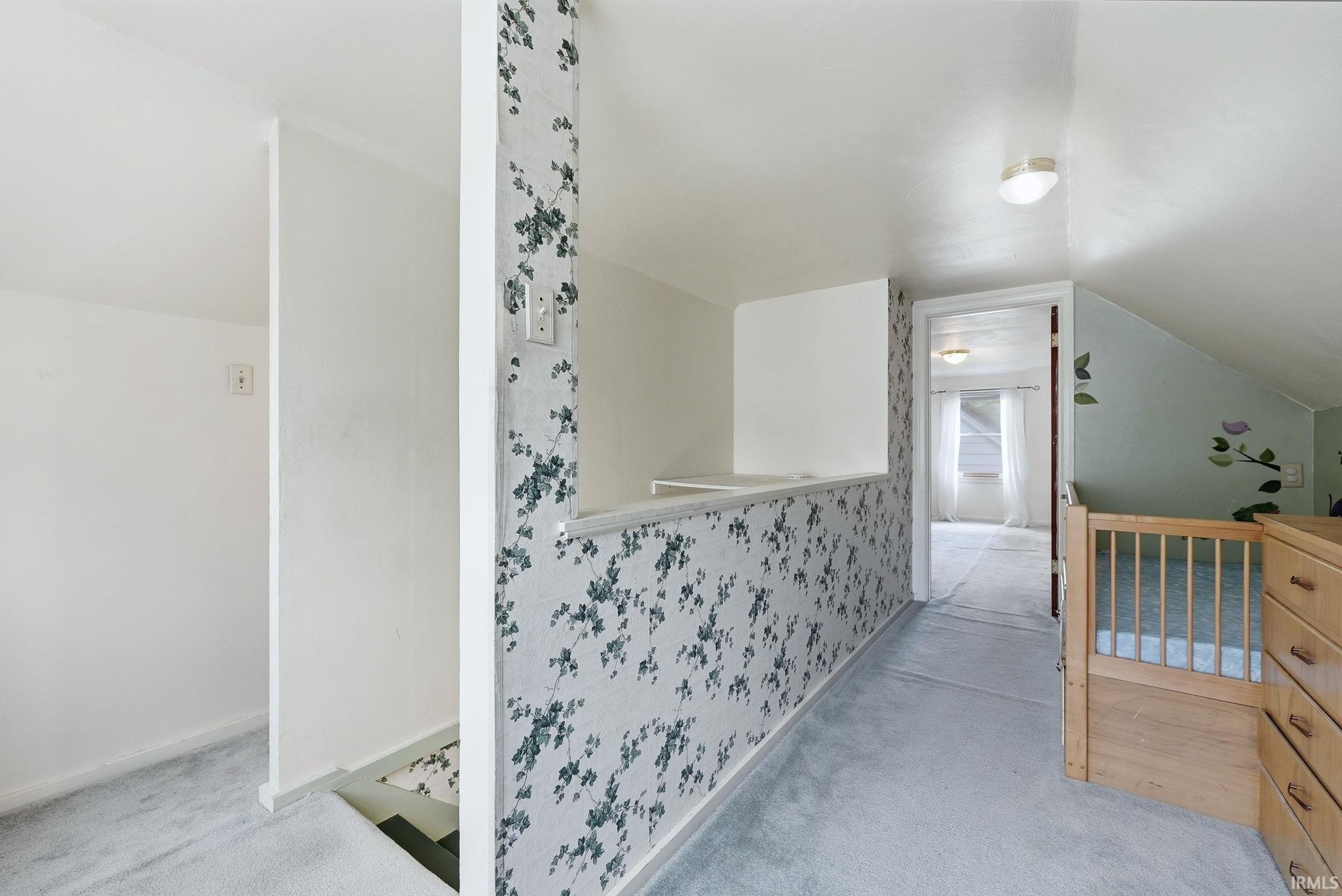 Carpeted hallway featuring a decorative half-wall with wallpaper, an overhead light fixture, and an open doorway leading to an additional carpeted room