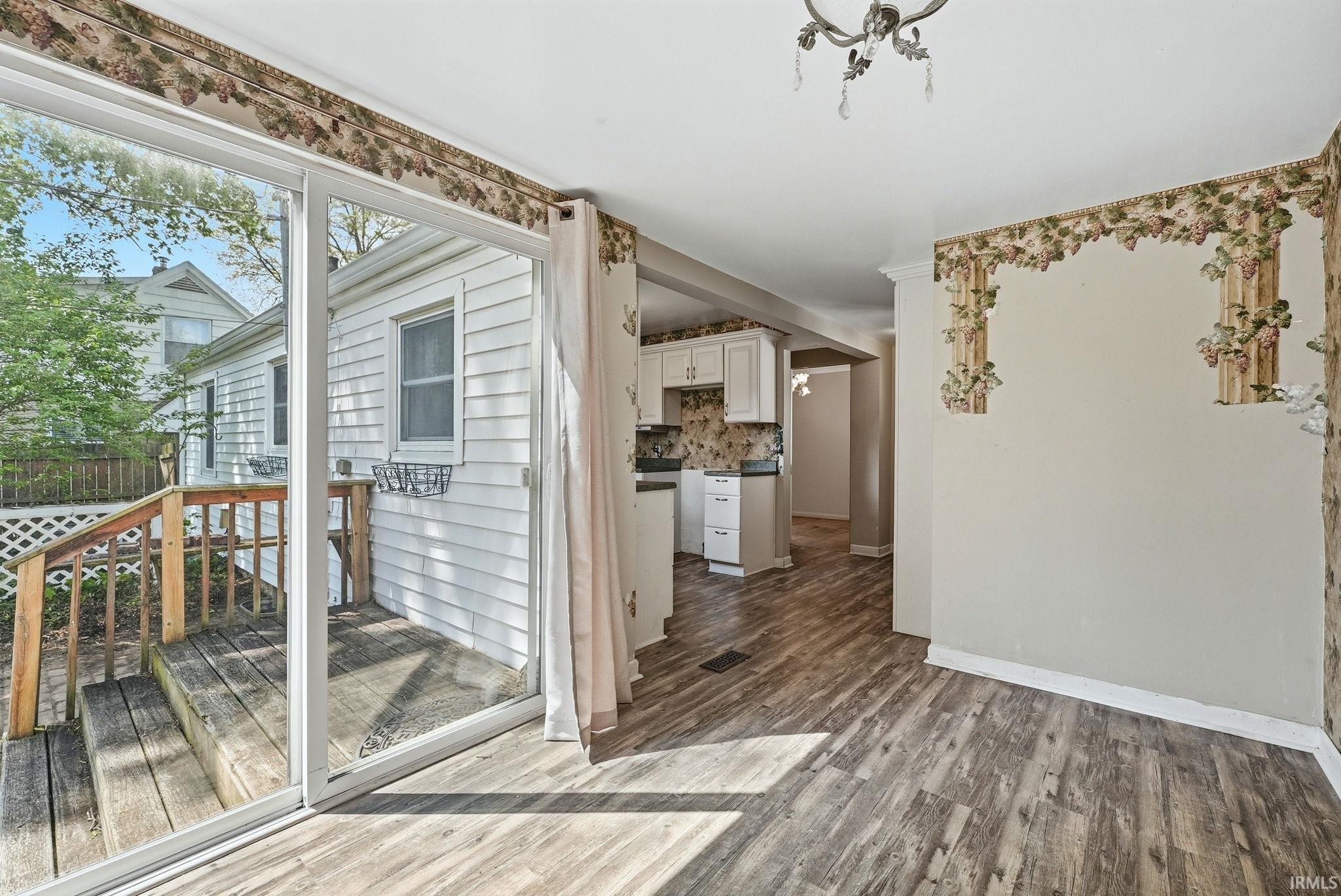 Spacious room with wood-finish flooring, sliding glass door access to an exterior wooden deck, and a view into a kitchen featuring white cabinetry