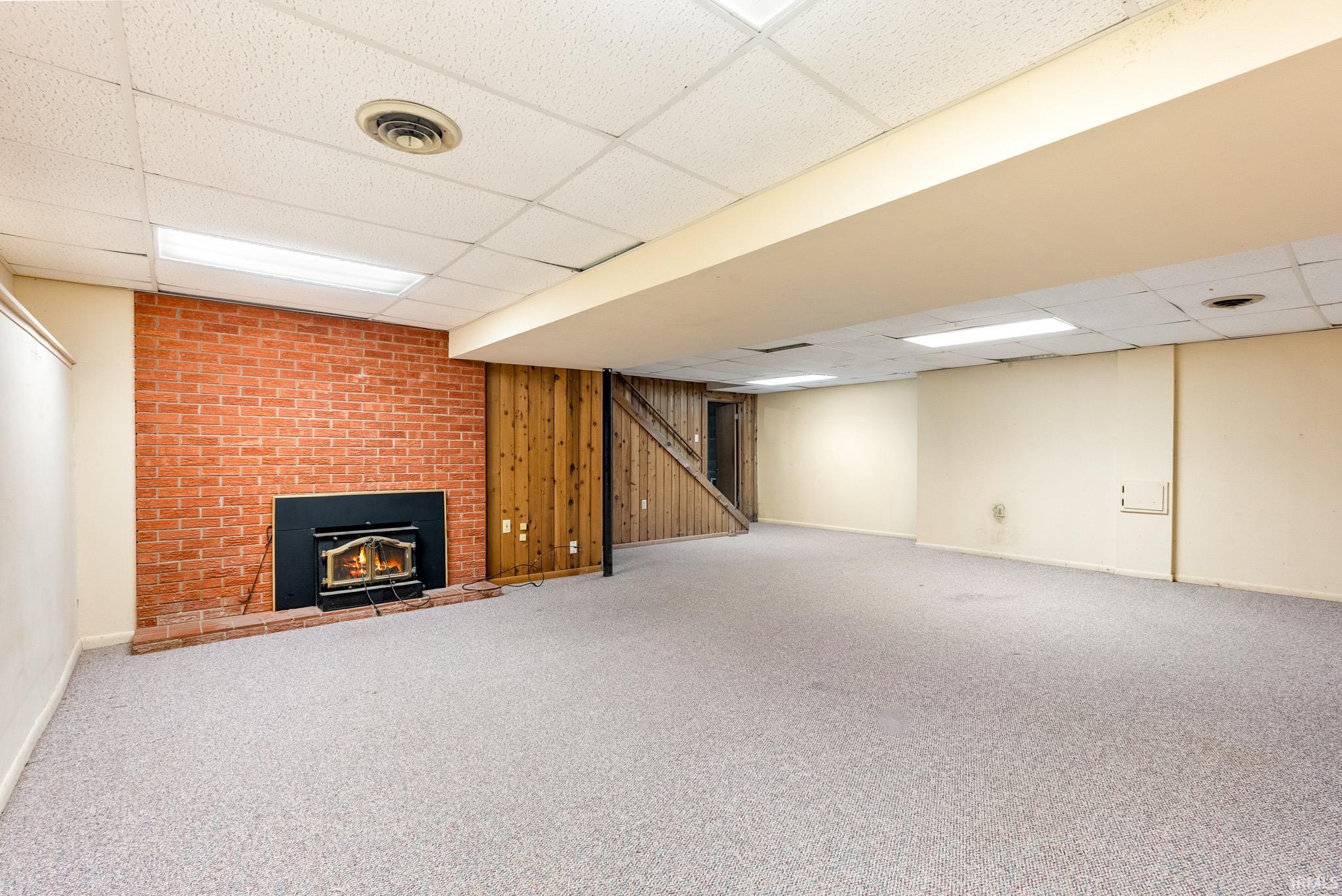 Spacious lower-level room featuring a brick-clad fireplace with an insert, wood-paneled accent wall, neutral carpeting, and a drop ceiling with integrated lighting