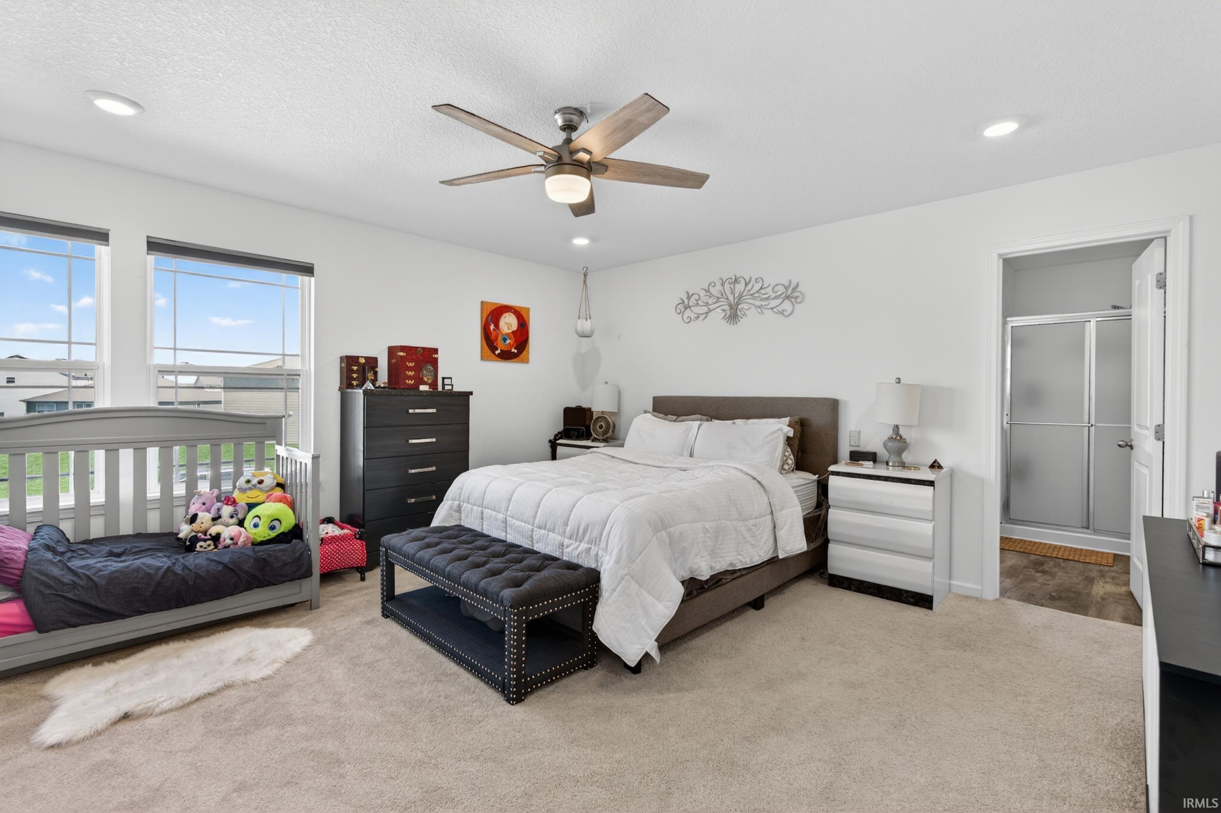 Carpeted bedroom with two windows, a ceiling fan with integrated lighting, recessed lighting, and an attached three-quarter bathroom with a sliding glass shower door