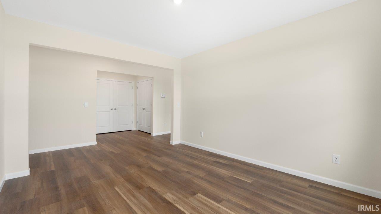 Spacious room featuring wood-finish flooring, light neutral wall paint, white baseboards, and a recessed doorway leading to two white paneled doors