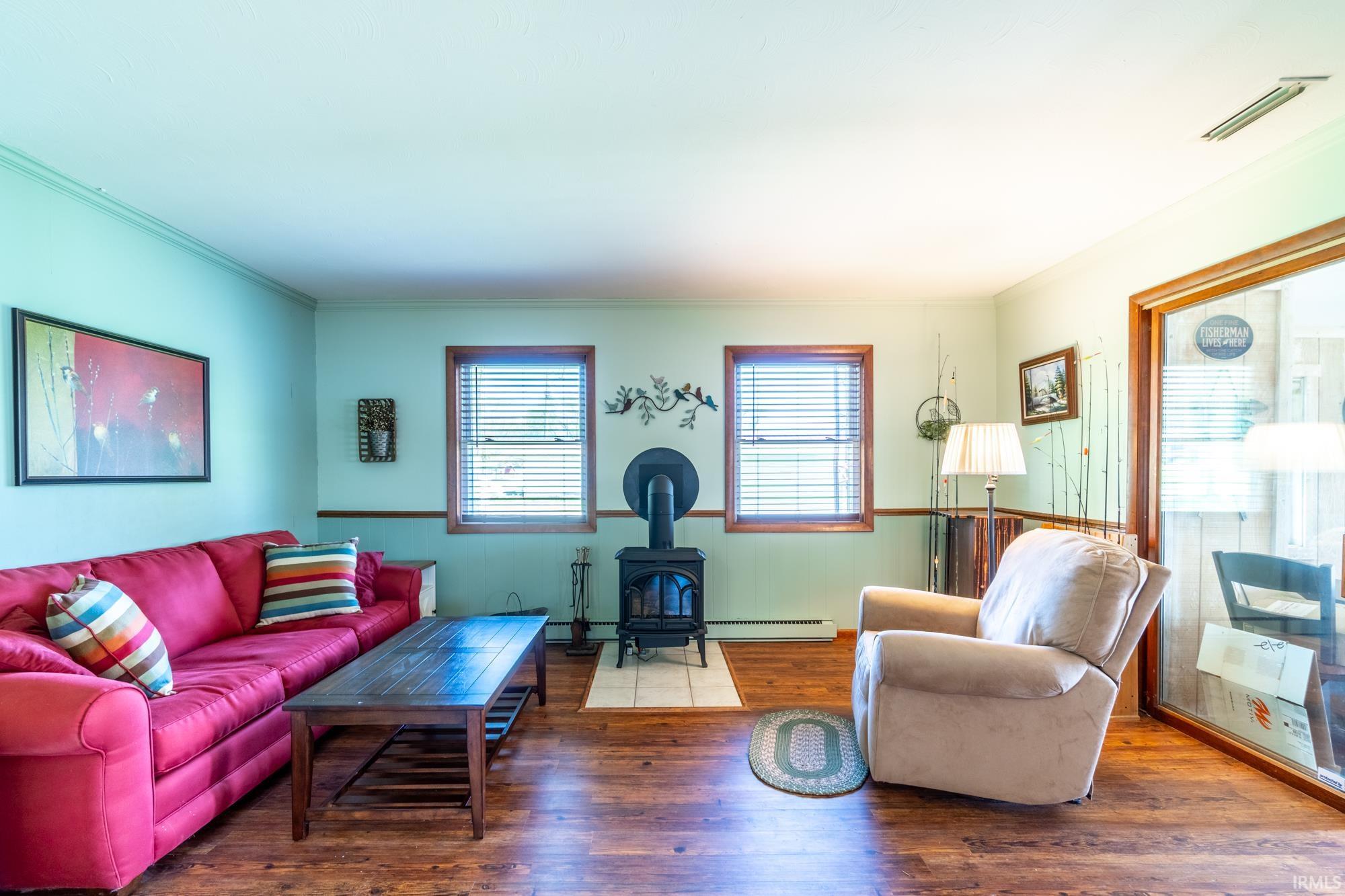 Living area featuring wood-finish flooring, two windows with blinds, and a wood-burning stove on a light-toned hearth