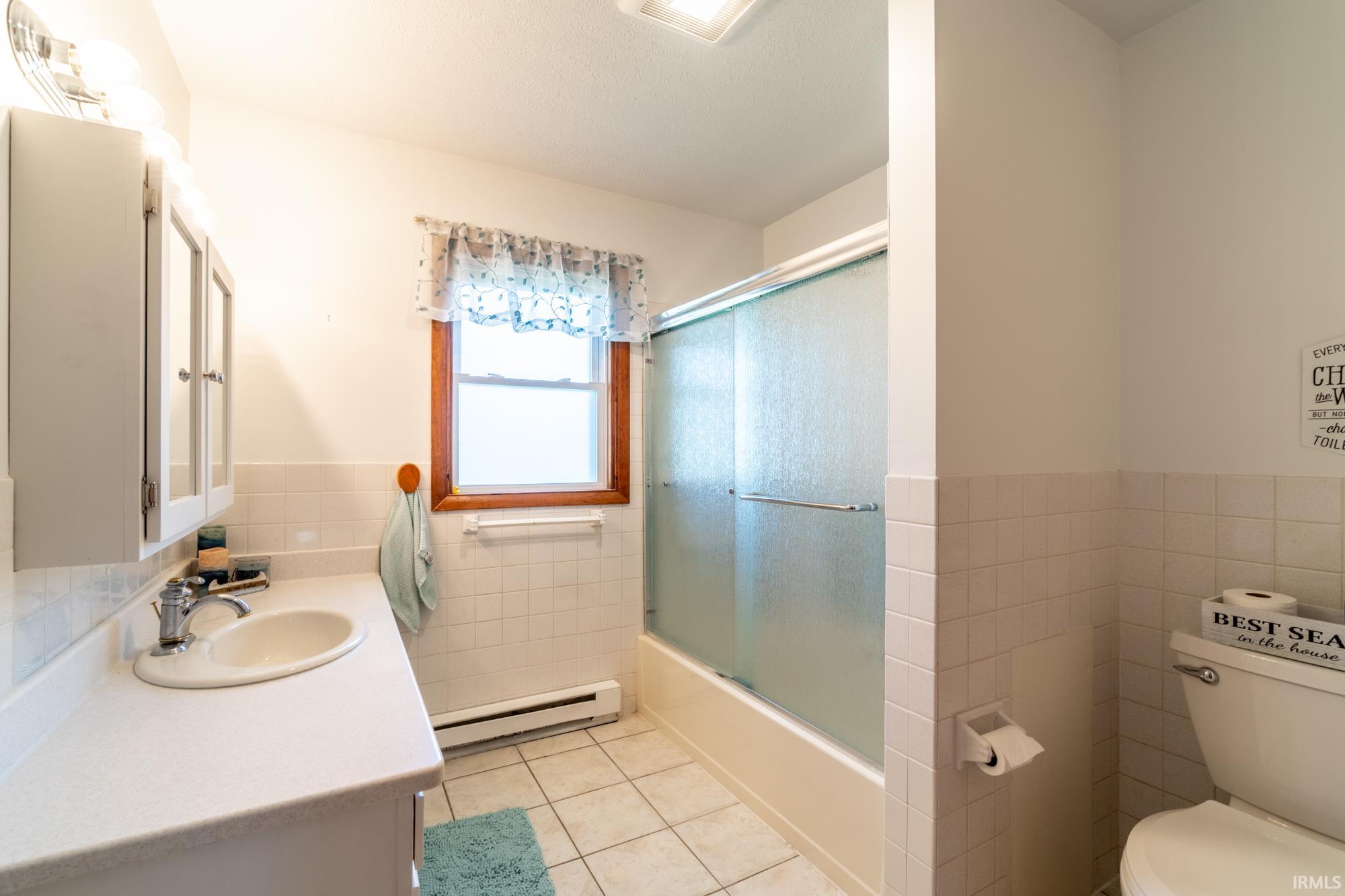 Bathroom featuring a vanity with an integrated sink, a medicine cabinet, tile flooring, a window with wood trim, and a bathtub with a sliding glass enclosure
