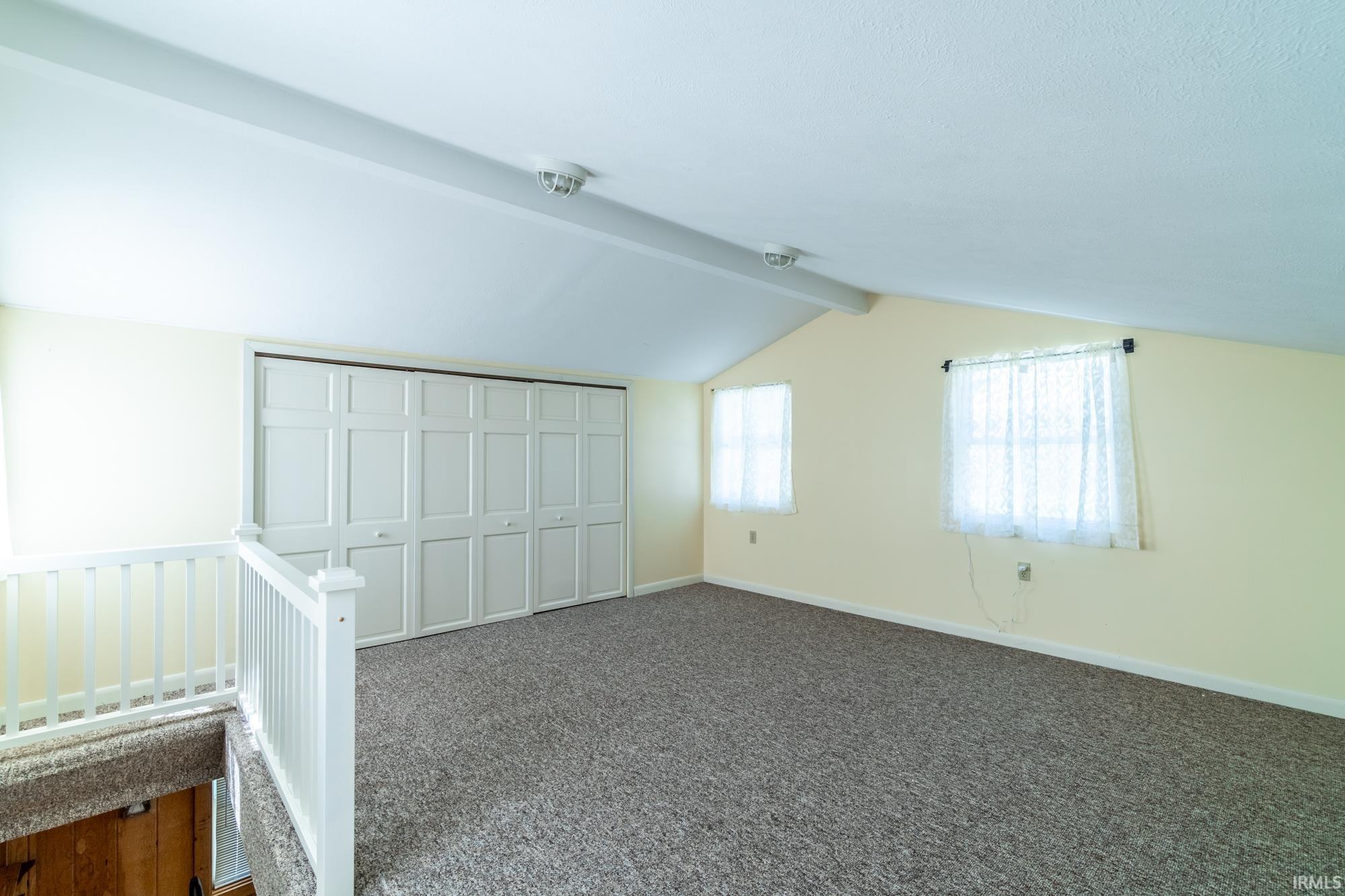 Carpeted loft area featuring a white railing, two windows, vaulted ceiling, and built-in closet with bi-fold doors