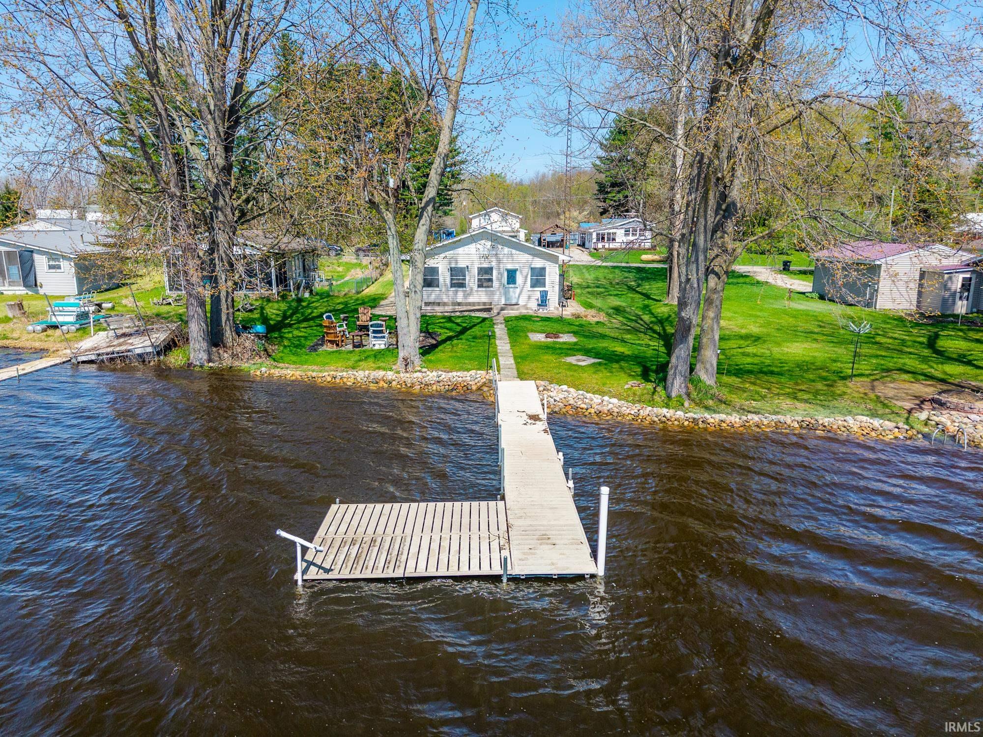Waterfront property featuring a private dock with an L-shaped extension, a gravel shoreline, and mature trees on a grassy lot