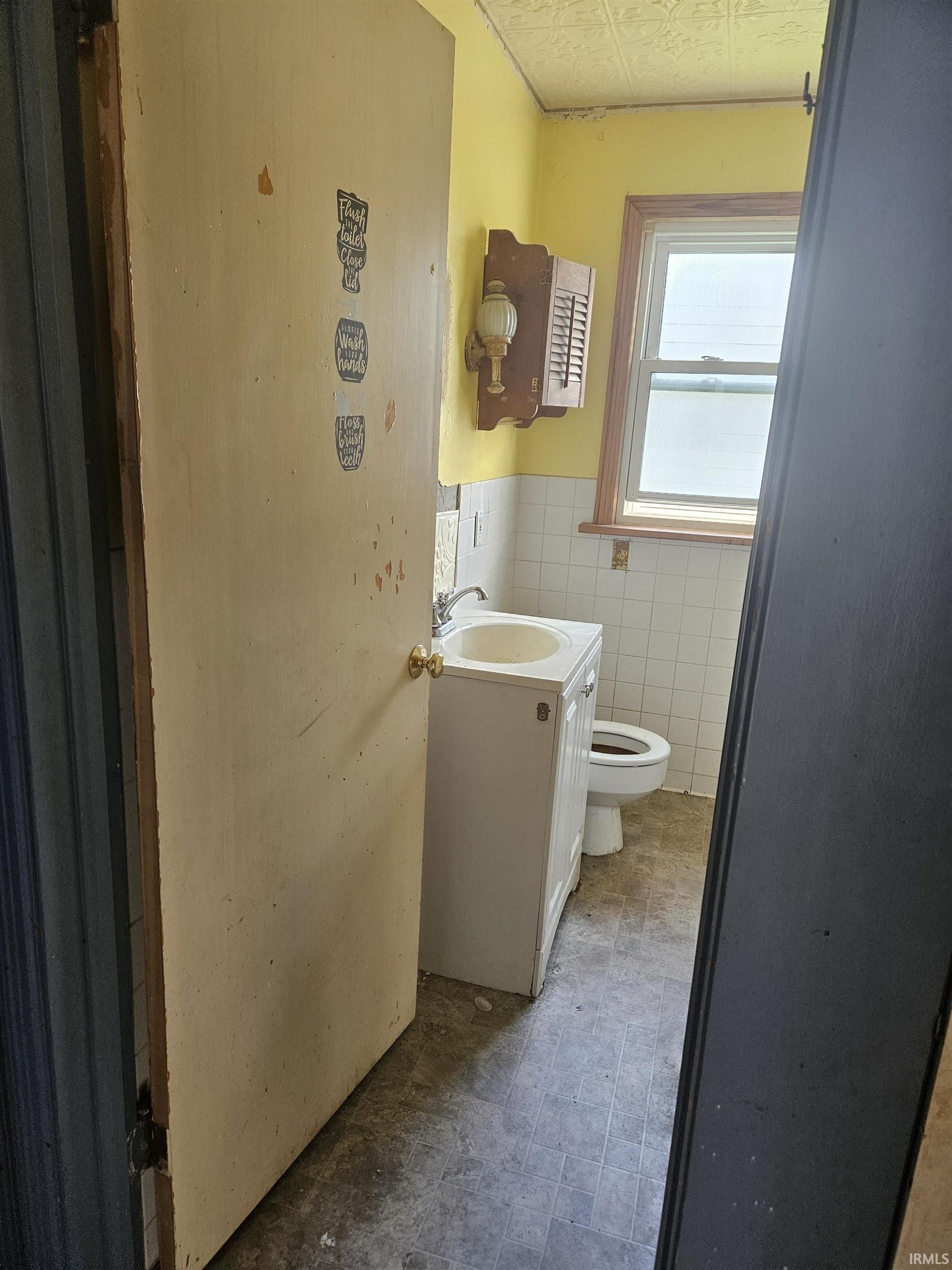 Bathroom featuring a single window with a wood frame, white ceramic wall tiling, a white vanity with an integrated sink, a commode, and a patterned ceiling