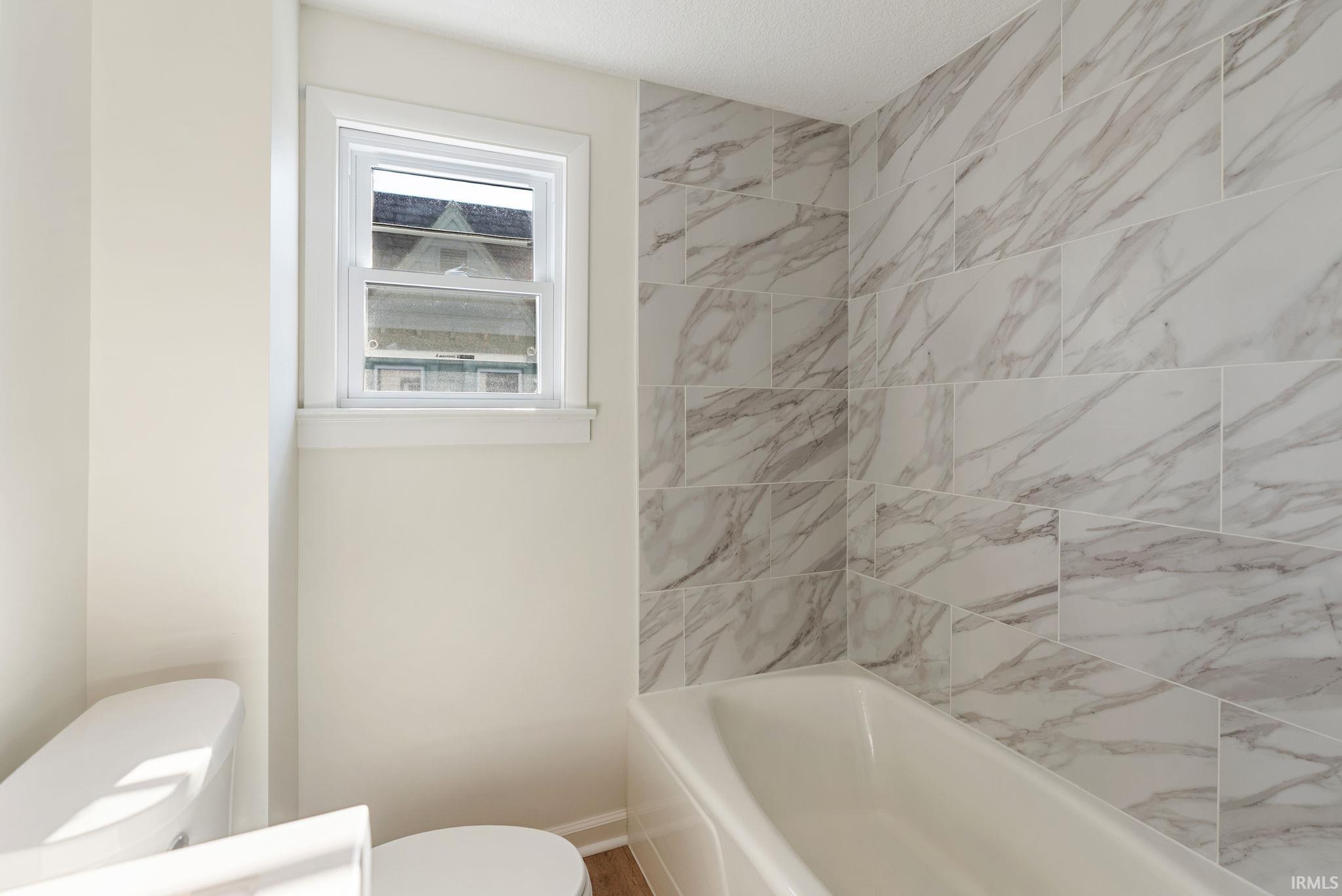 Bathroom featuring a white bathtub, a toilet, a window with white trim, and marble-look tile surrounding the tub