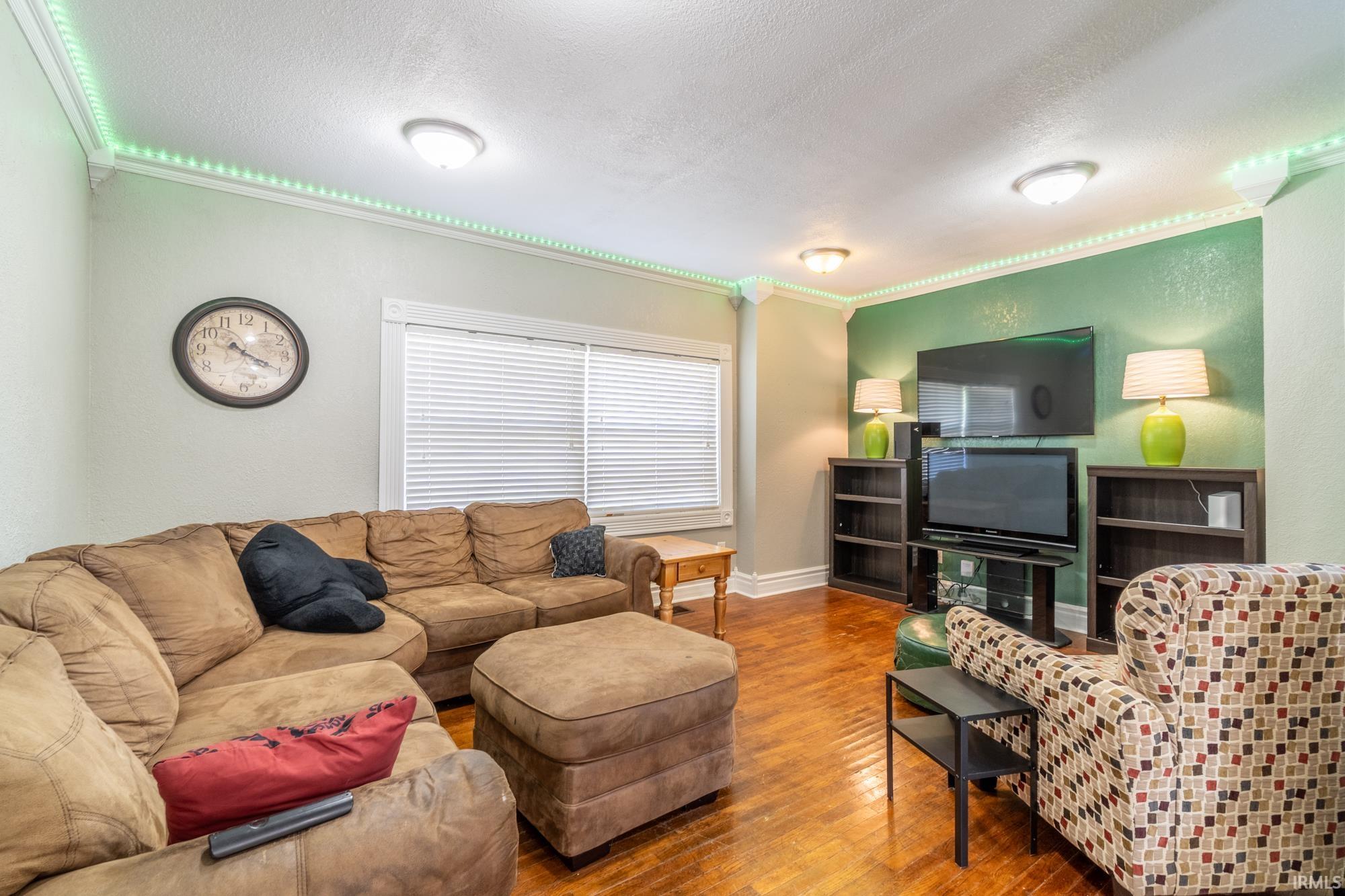 Living area featuring wood-finish flooring, crown molding with accent lighting, and a window with horizontal blinds