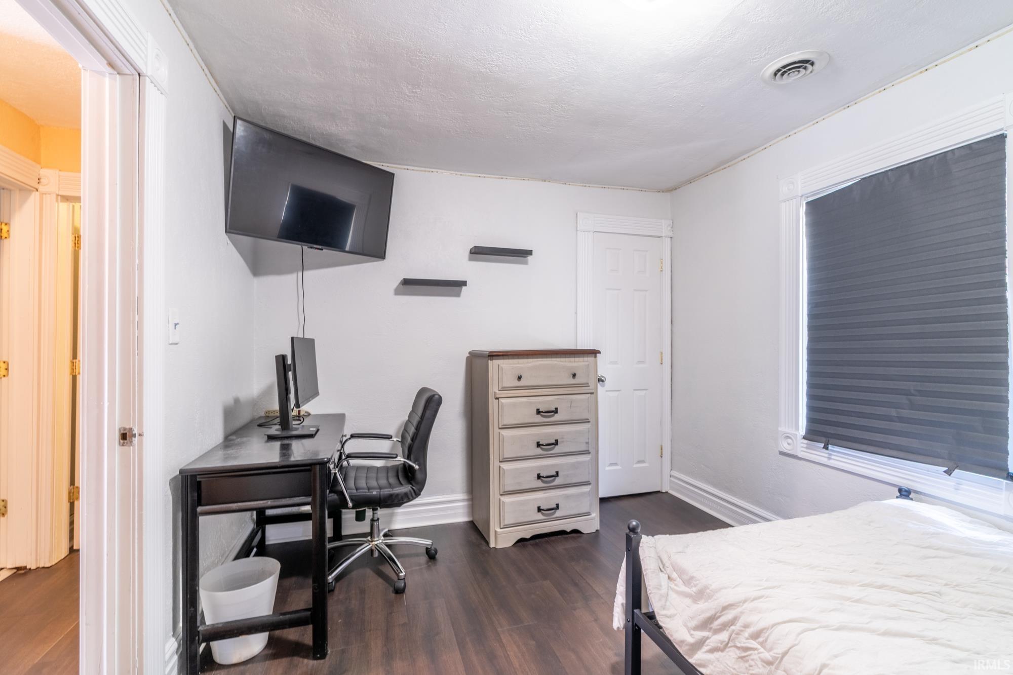 Interior room featuring dark wood-finish flooring, white walls, and a window with a pleated shade