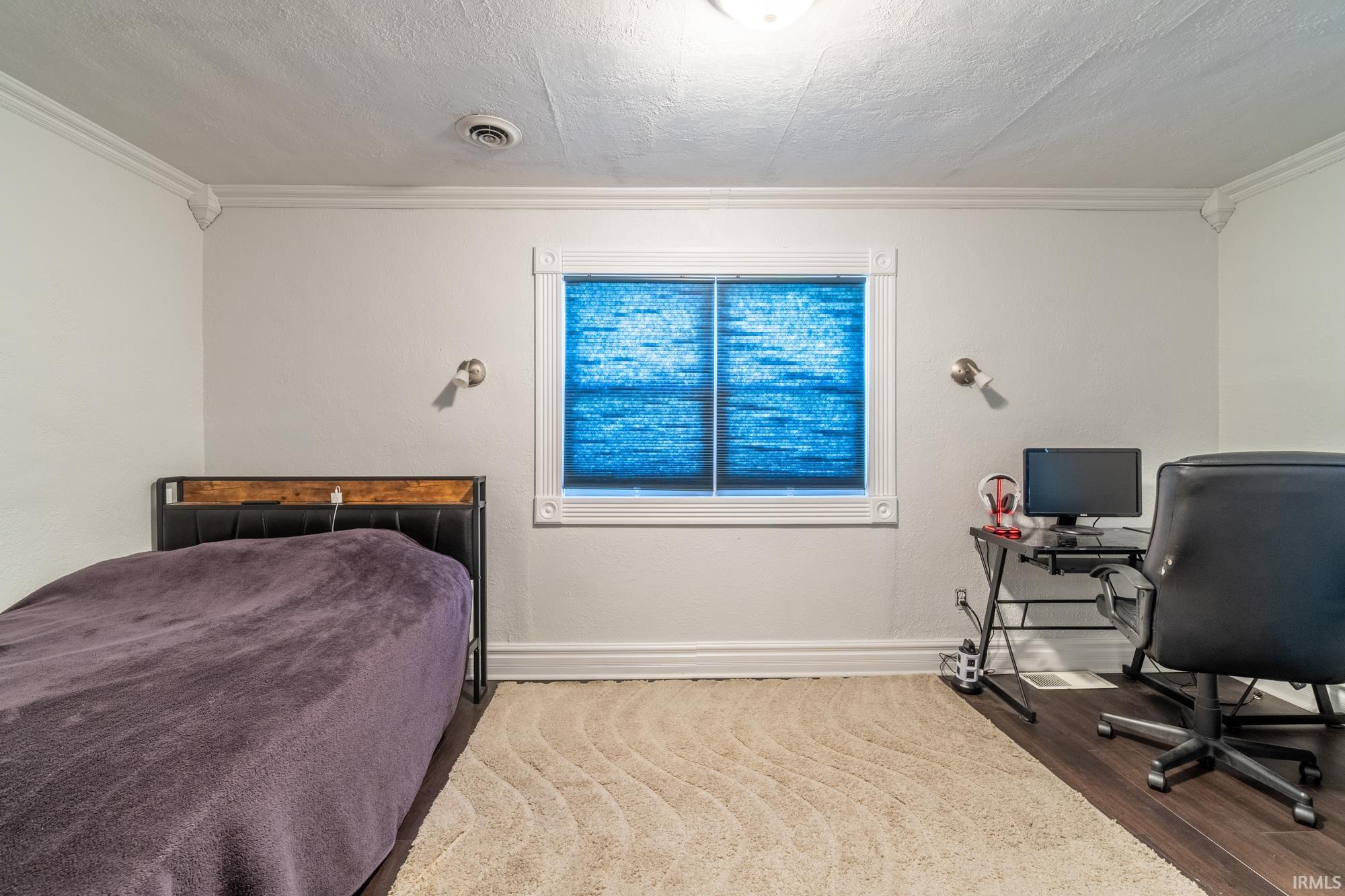 Bedroom featuring crown molding, wall-mounted sconces, and a window with custom blinds