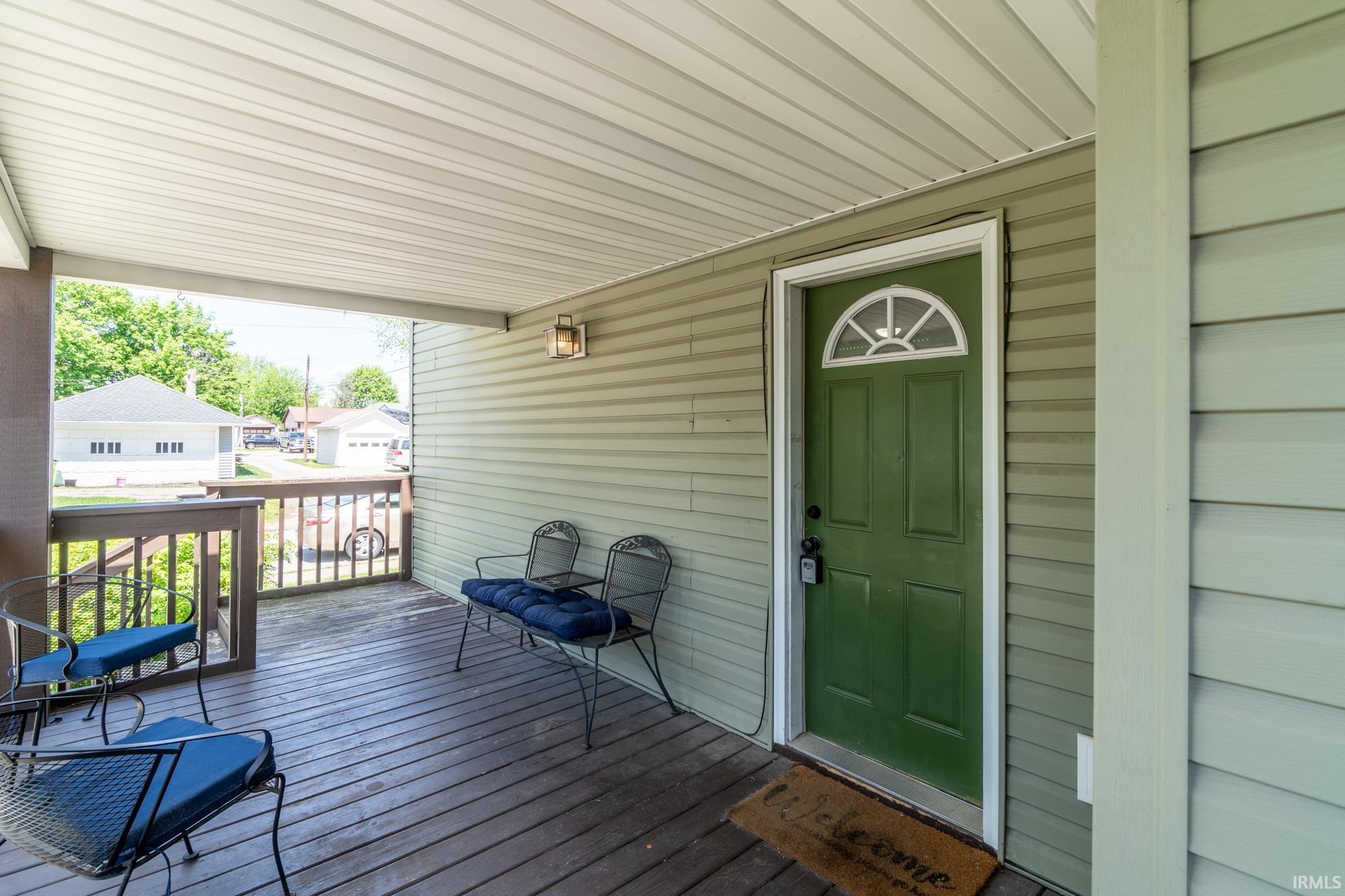 Covered porch with wood-finish flooring, a green entry door featuring an arched window, light-colored siding, and a wooden railing