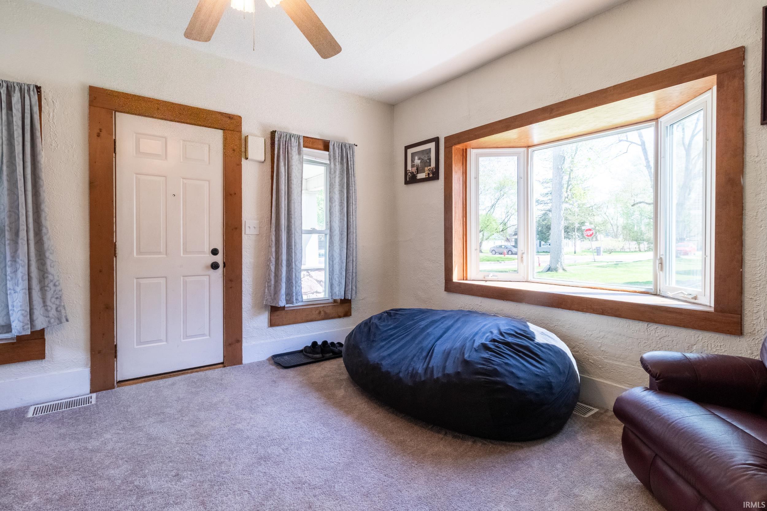 Living area featuring a spacious bay window with wood trim, light gray carpeting, and a ceiling fan